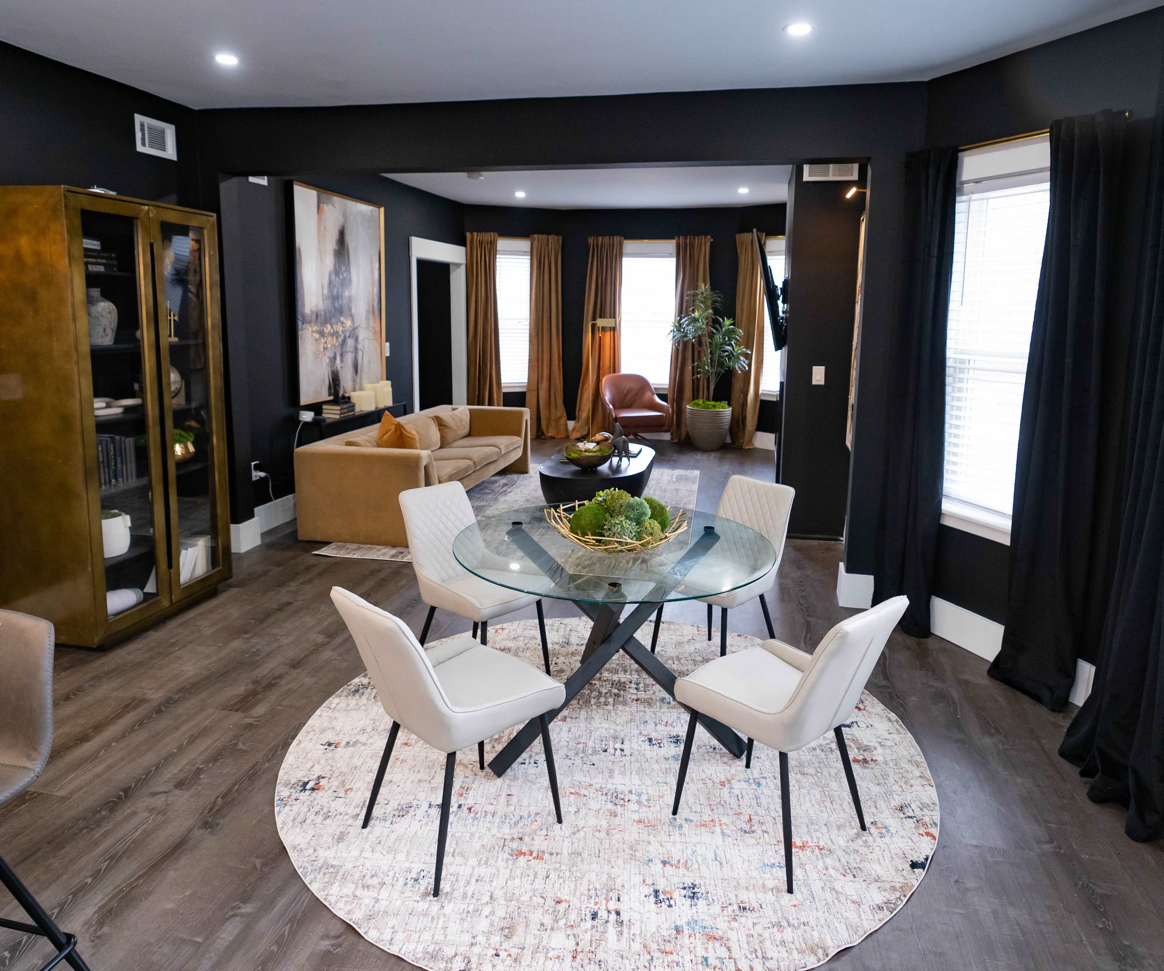 A dining area features a glass table surrounded by four chairs on a patterned rug, with a living space and large windows in the background.
