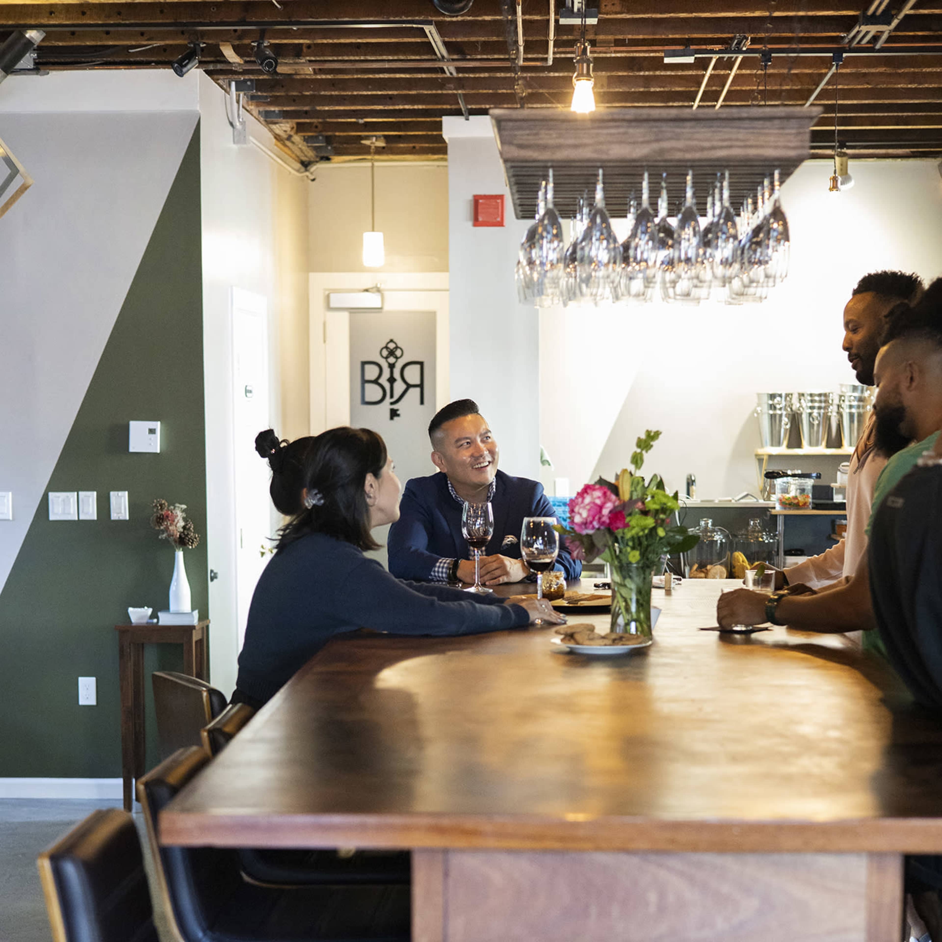 A group of people are seated around a wooden bar in a modern space, engaged in conversation while drinks and flowers are placed on the counter.