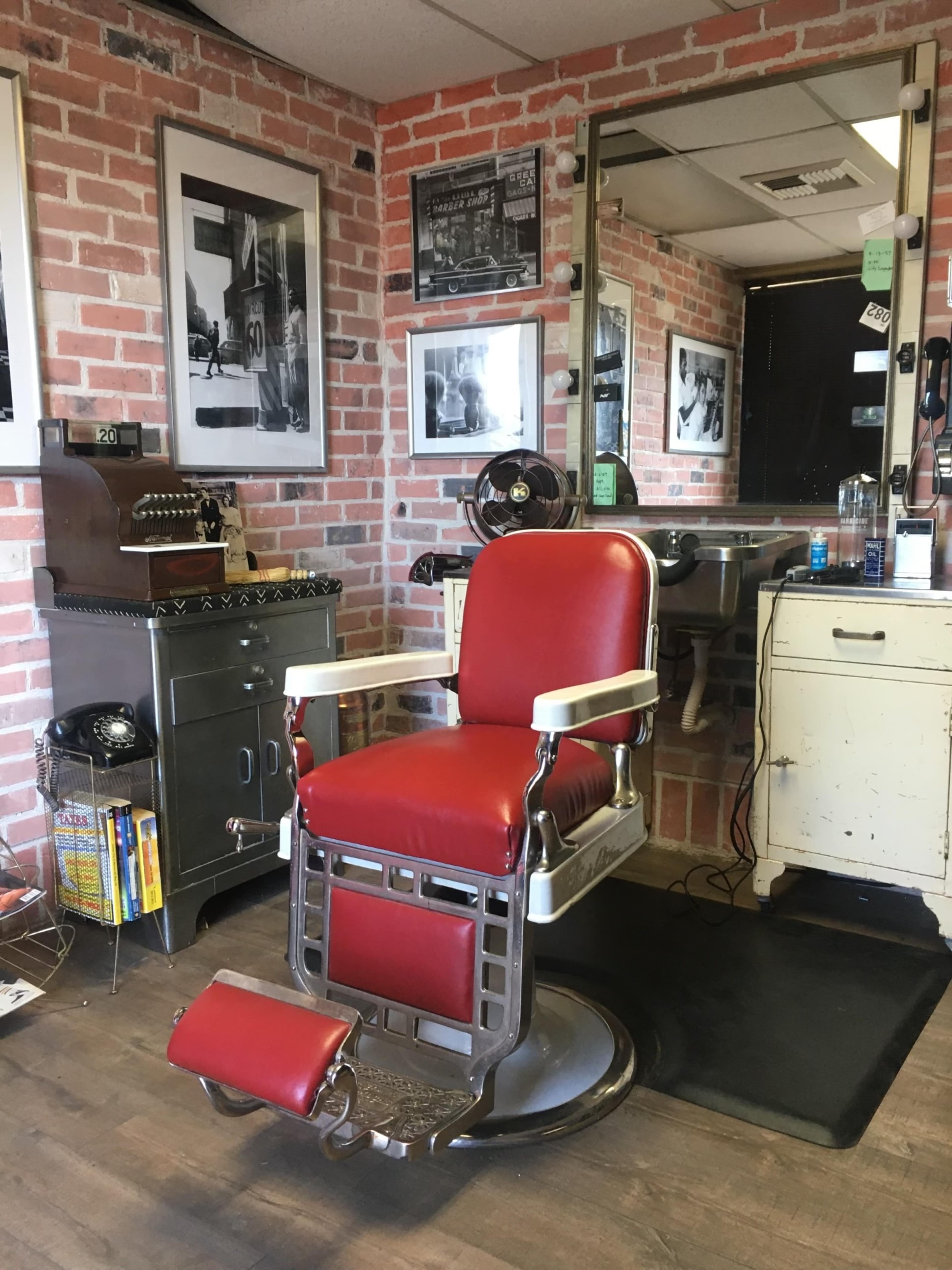A vintage barber shop interior featuring a red barber chair, brick walls adorned with black and white photographs, and various tools and equipment for hair cutting.