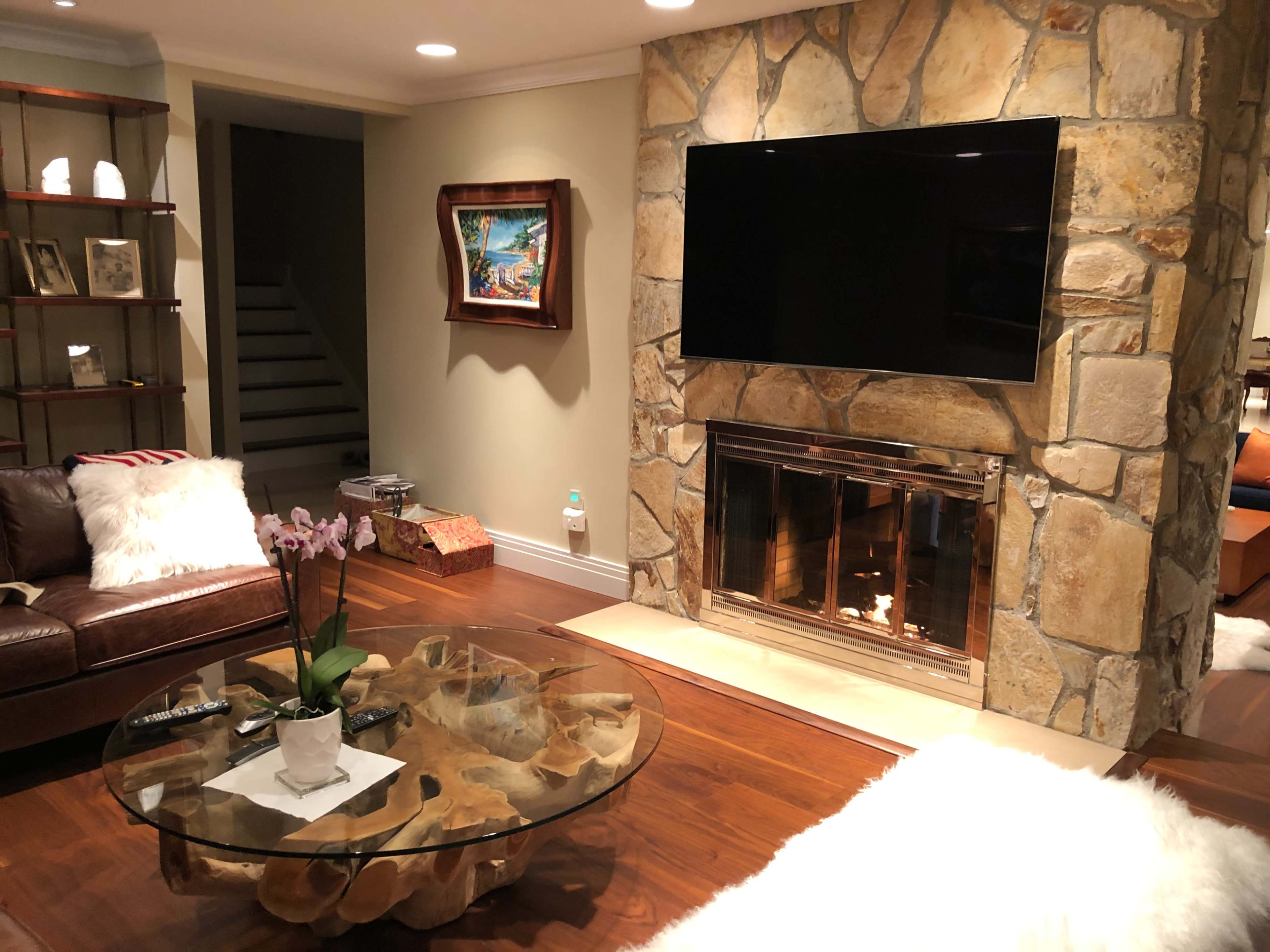 The image shows a living room with a stone fireplace, a large flat-screen television mounted above it, a glass coffee table, and a leather sofa, with stairs visible in the background.