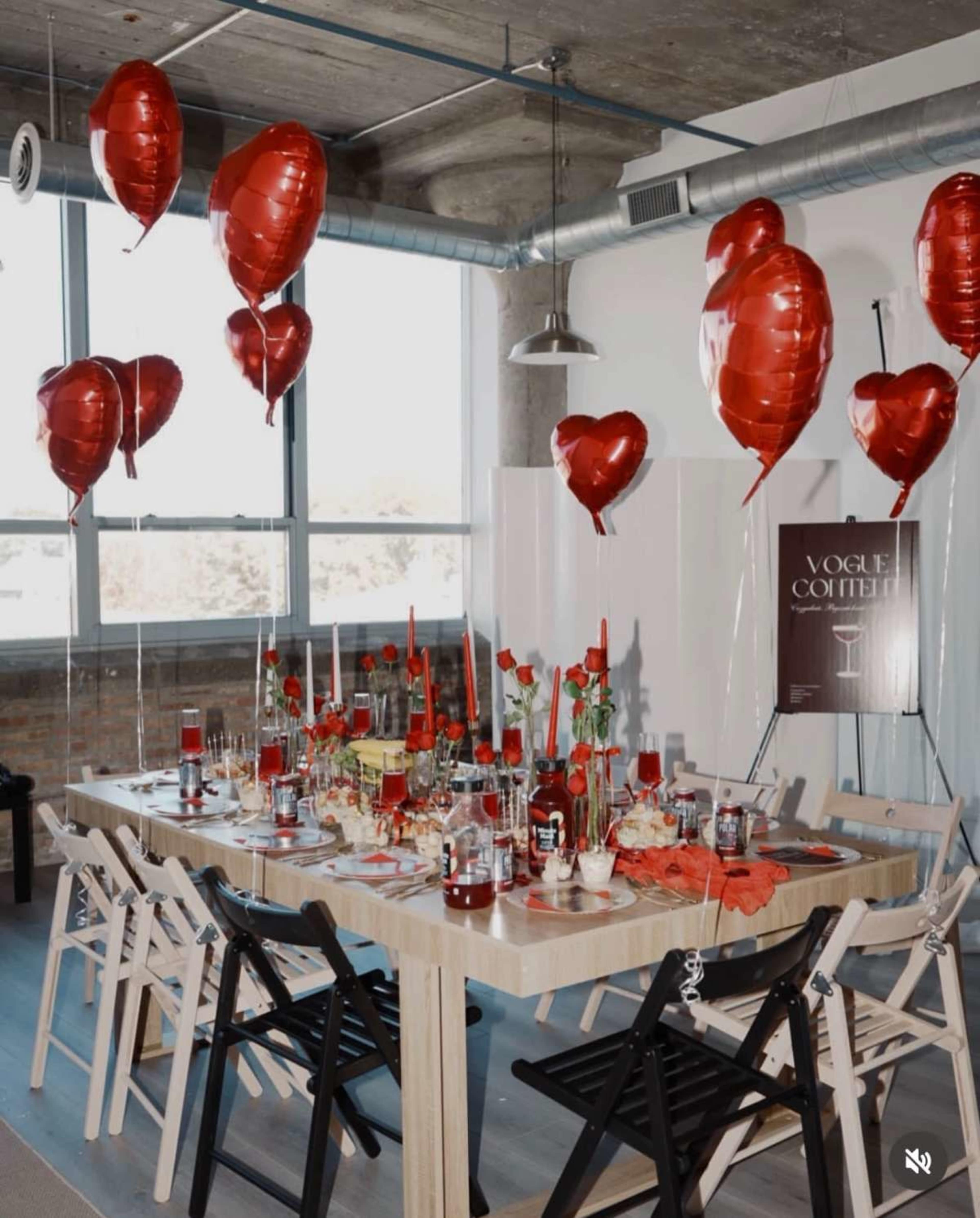 A dining table is adorned with red balloons, roses, and decorative items, set against a backdrop of large windows.