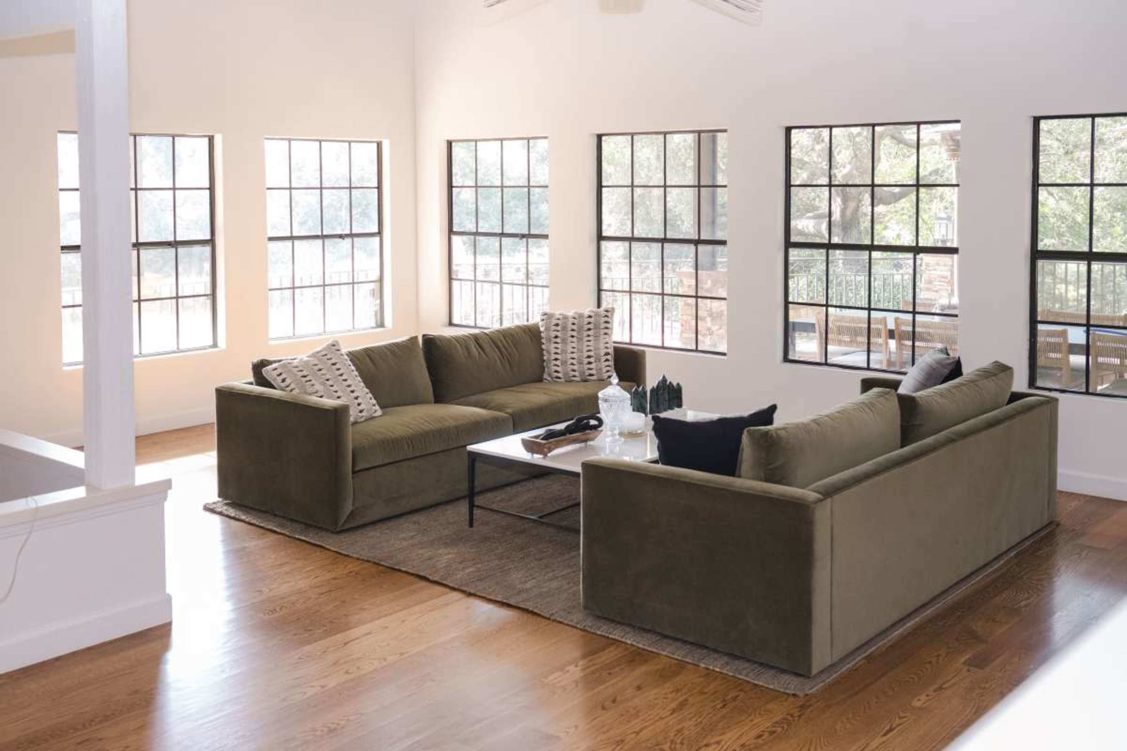 The image shows a spacious living room with a large sectional sofa, a coffee table, and multiple windows allowing natural light to fill the space.
