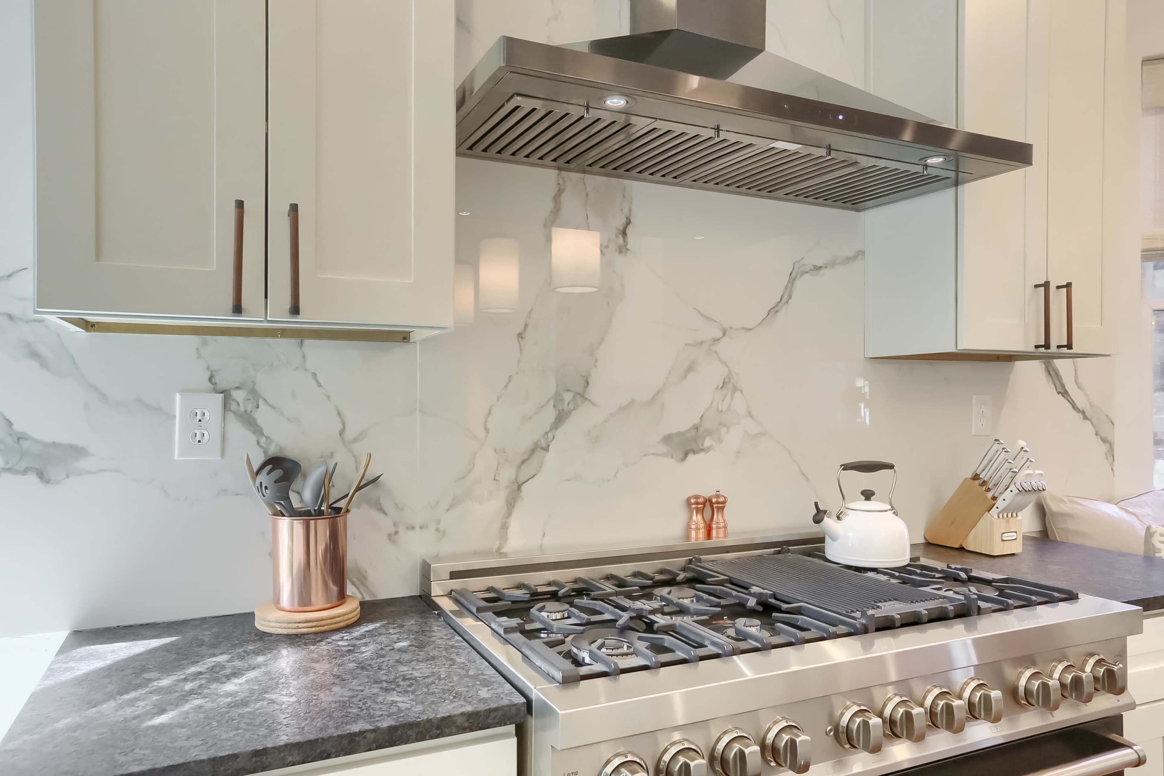 A modern kitchen featuring a gas range with a stainless steel hood, marble backsplash, and a copper utensil holder on the counter.