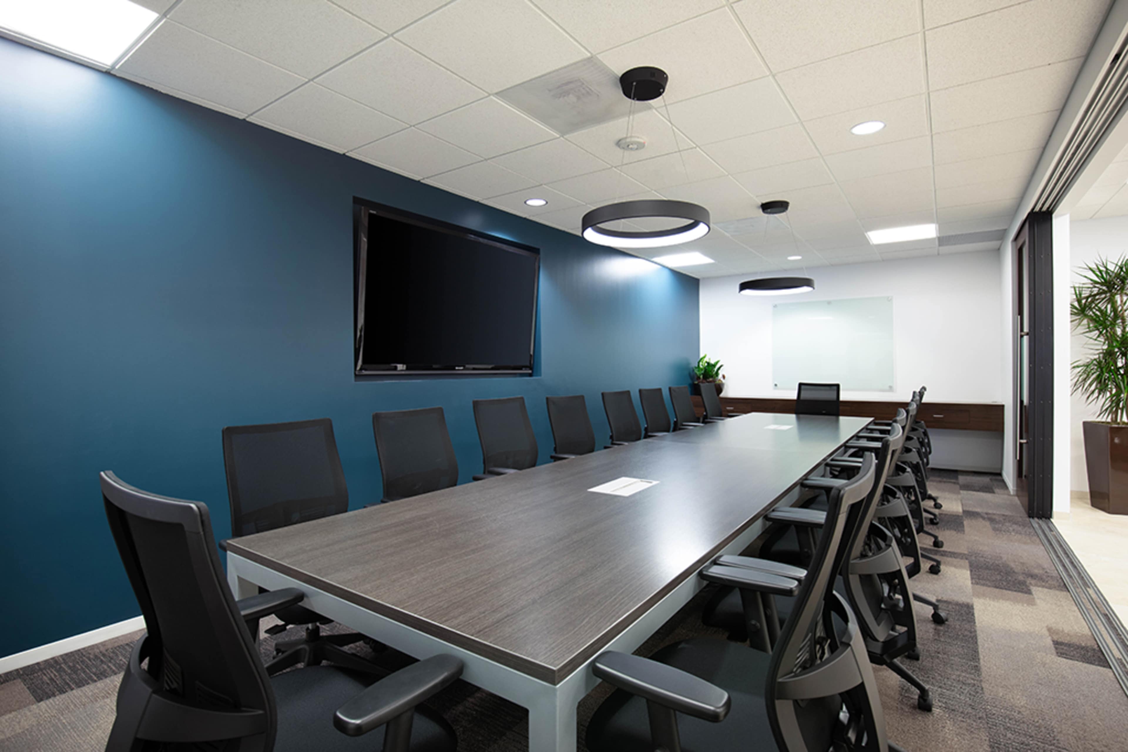 A modern conference room features a long table surrounded by black chairs, with a large television mounted on the wall and a whiteboard in the background.