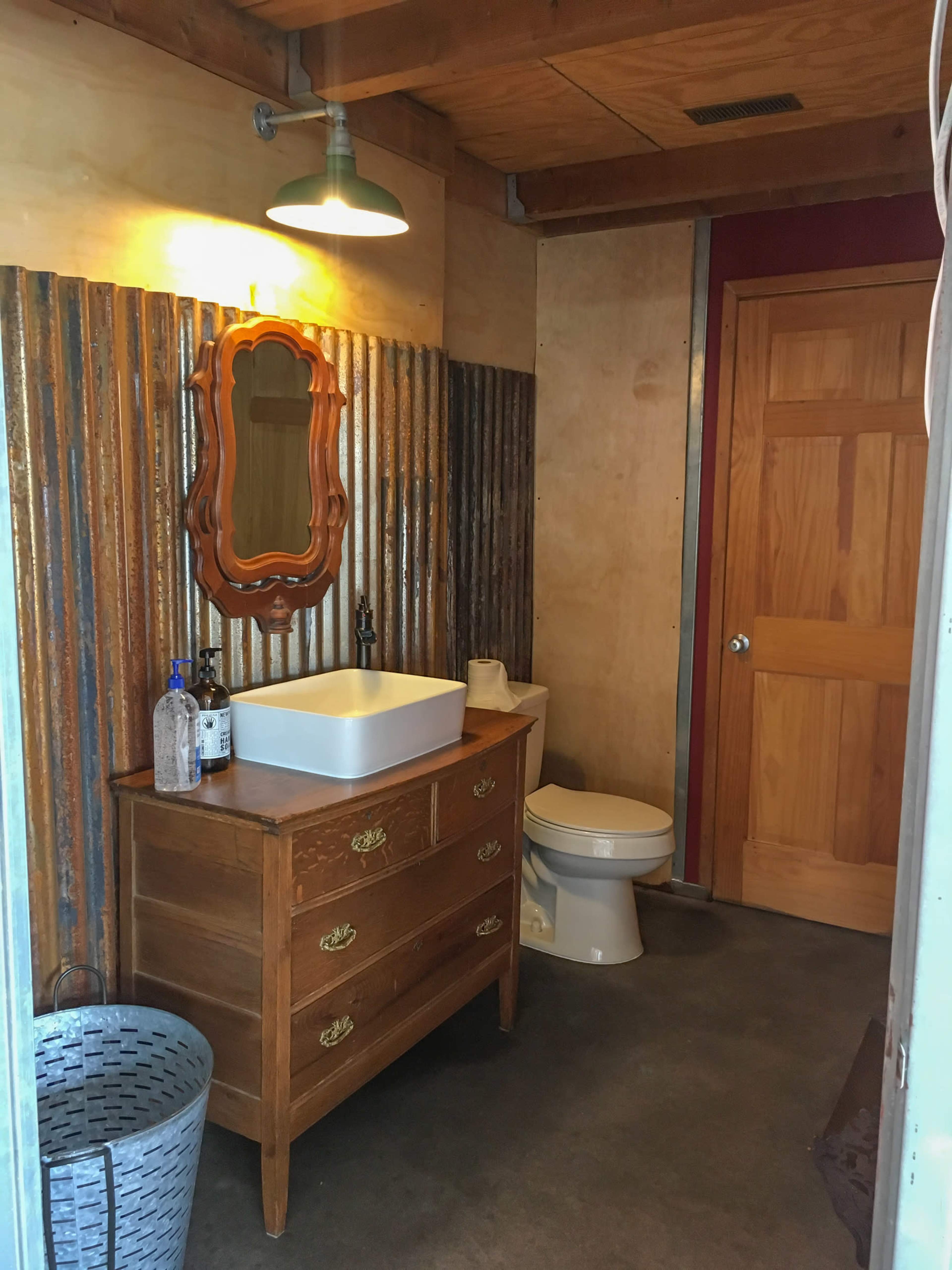 A rustic bathroom featuring a wooden dresser with a sink, a toilet, and a wall made of corrugated metal.