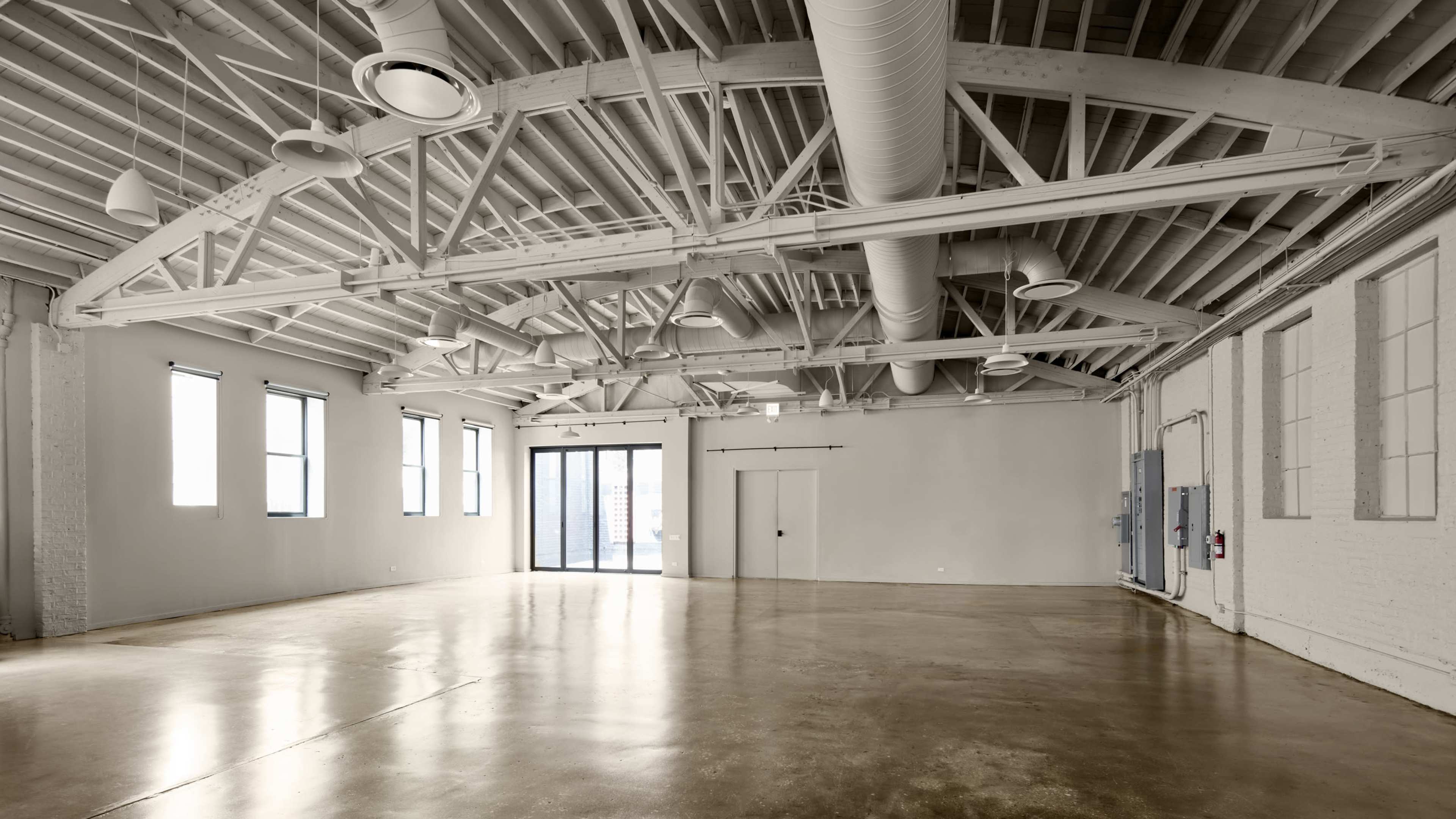 The image shows an empty, spacious industrial-style room with tall ceilings, exposed beams, and large windows.
