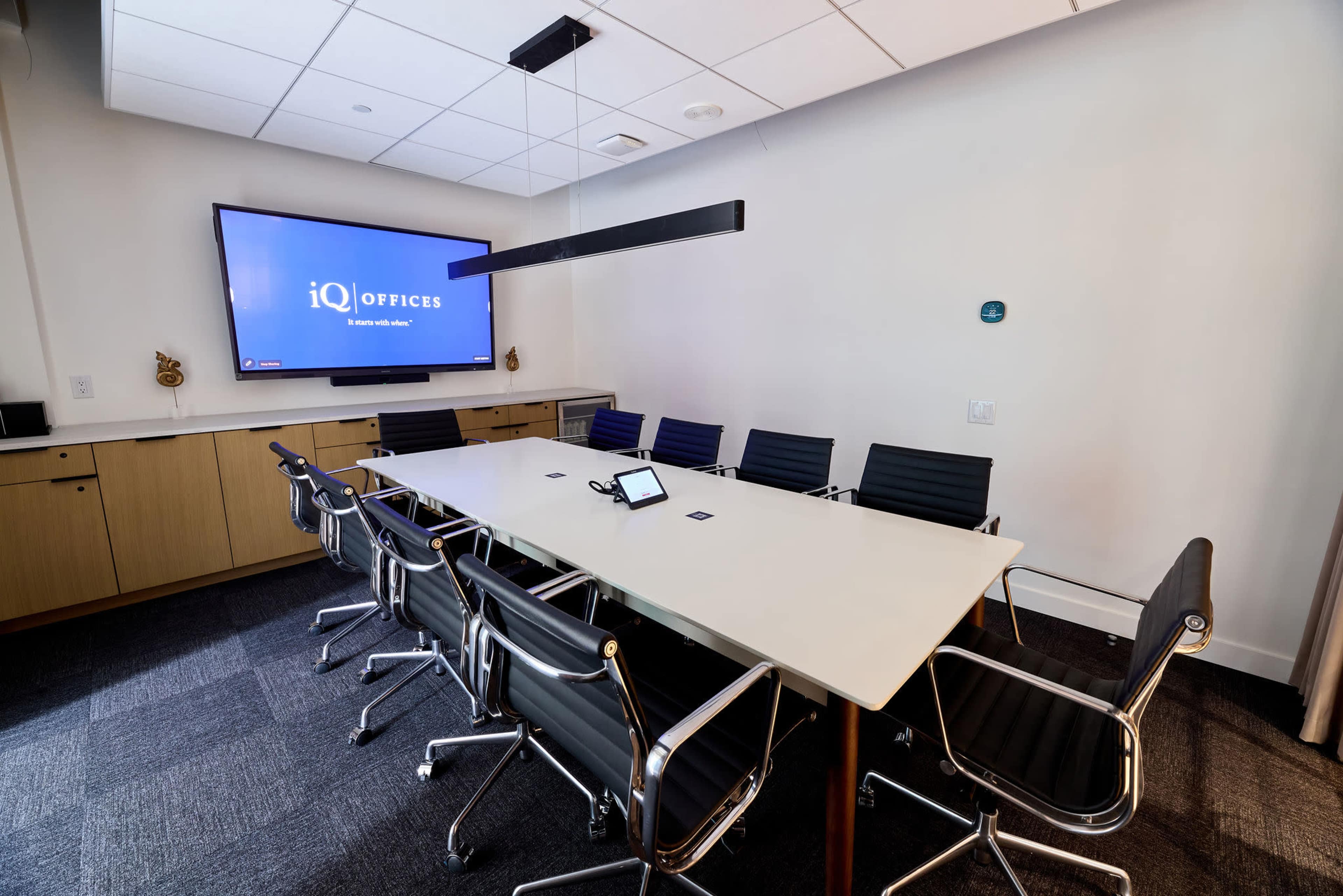 A modern conference room features a large table surrounded by sleek chairs, with a television screen displaying the iQ Offices logo on the wall.