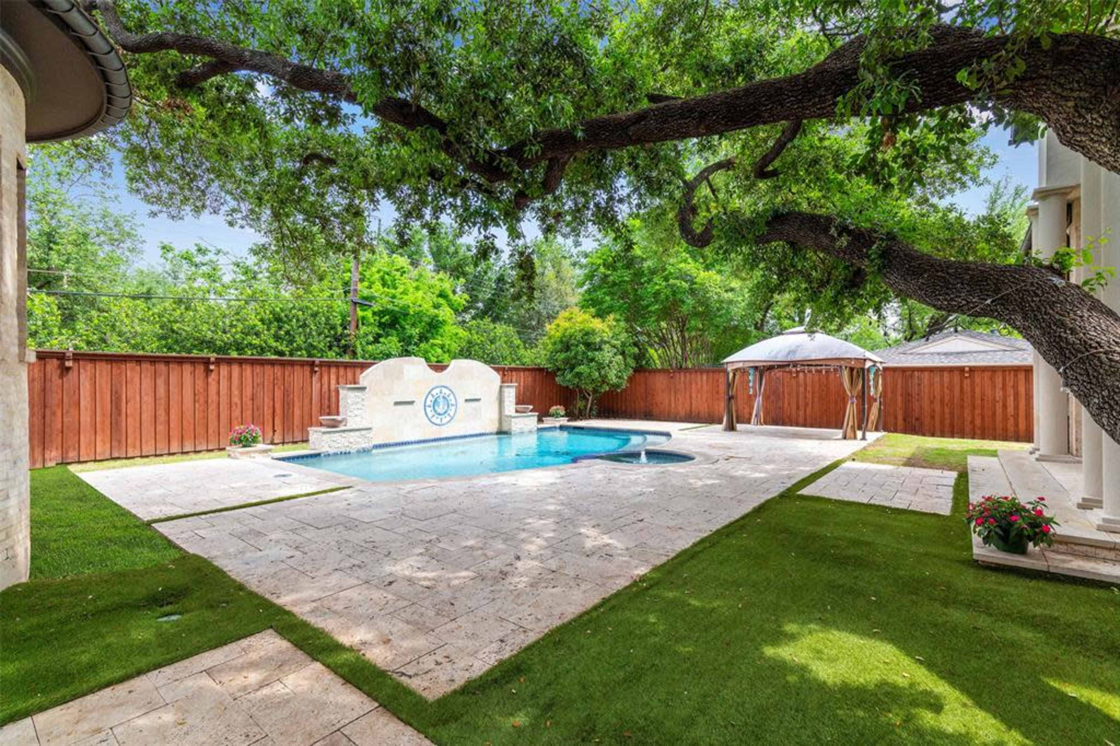 The image shows a swimming pool surrounded by stone tiles and grass, with a structured wall feature and two canopies in the background.