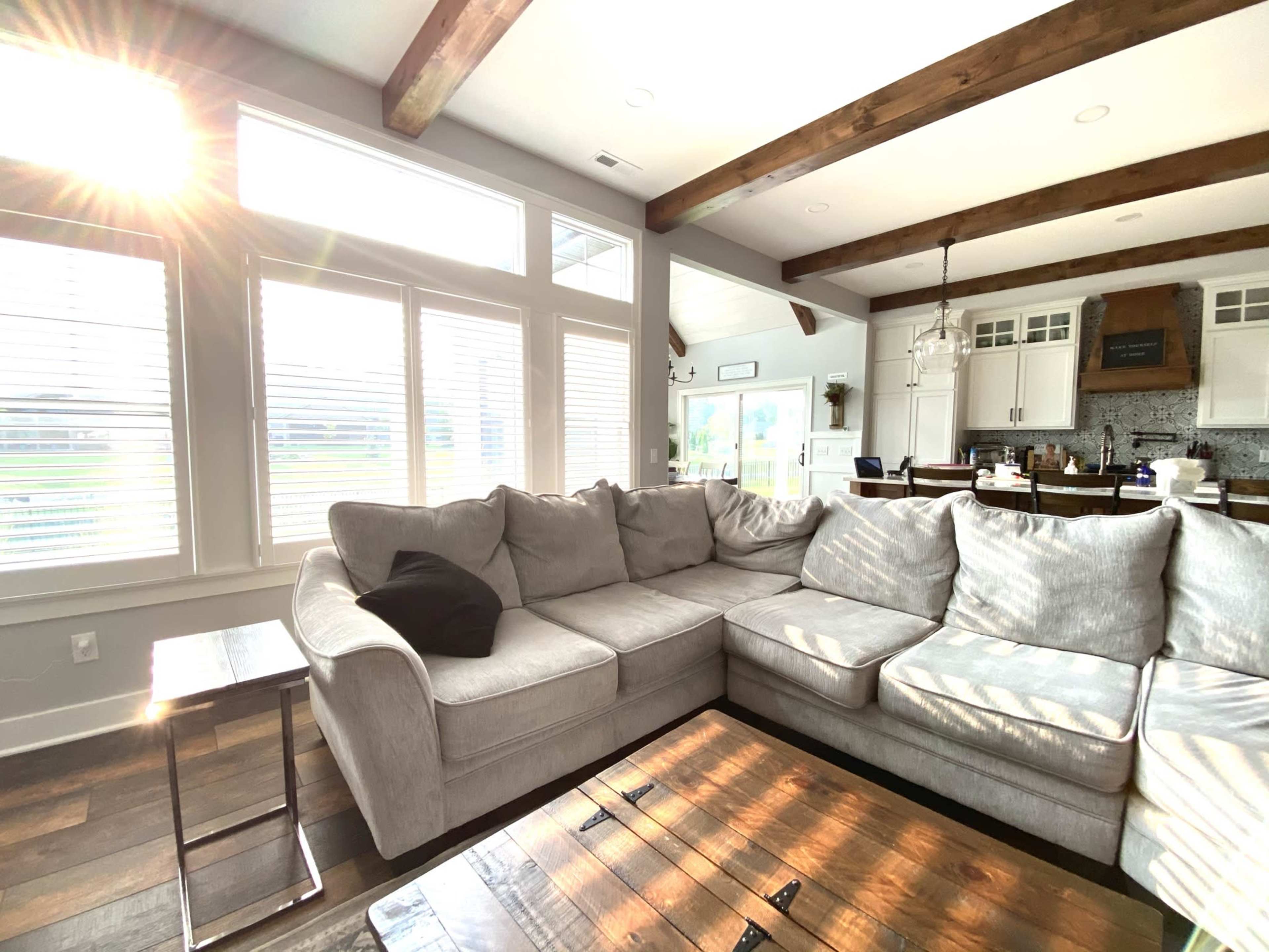 A spacious living room features a large sectional sofa, a wooden coffee table, and sunlight streaming through multiple windows.
