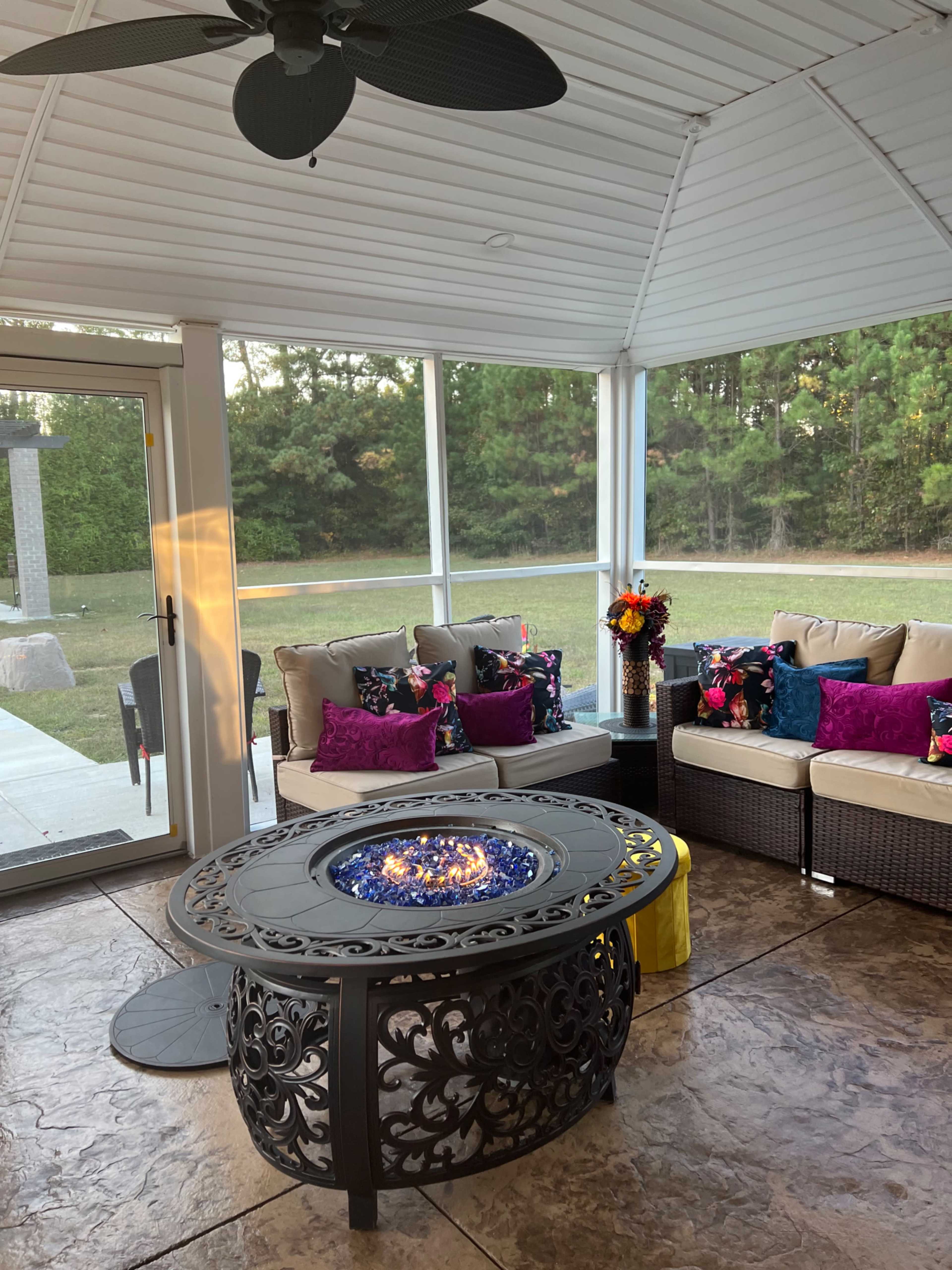 A screened porch with a decorative fire table in the center, surrounded by a sectional sofa adorned with colorful cushions and a view of a grassy area beyond.
