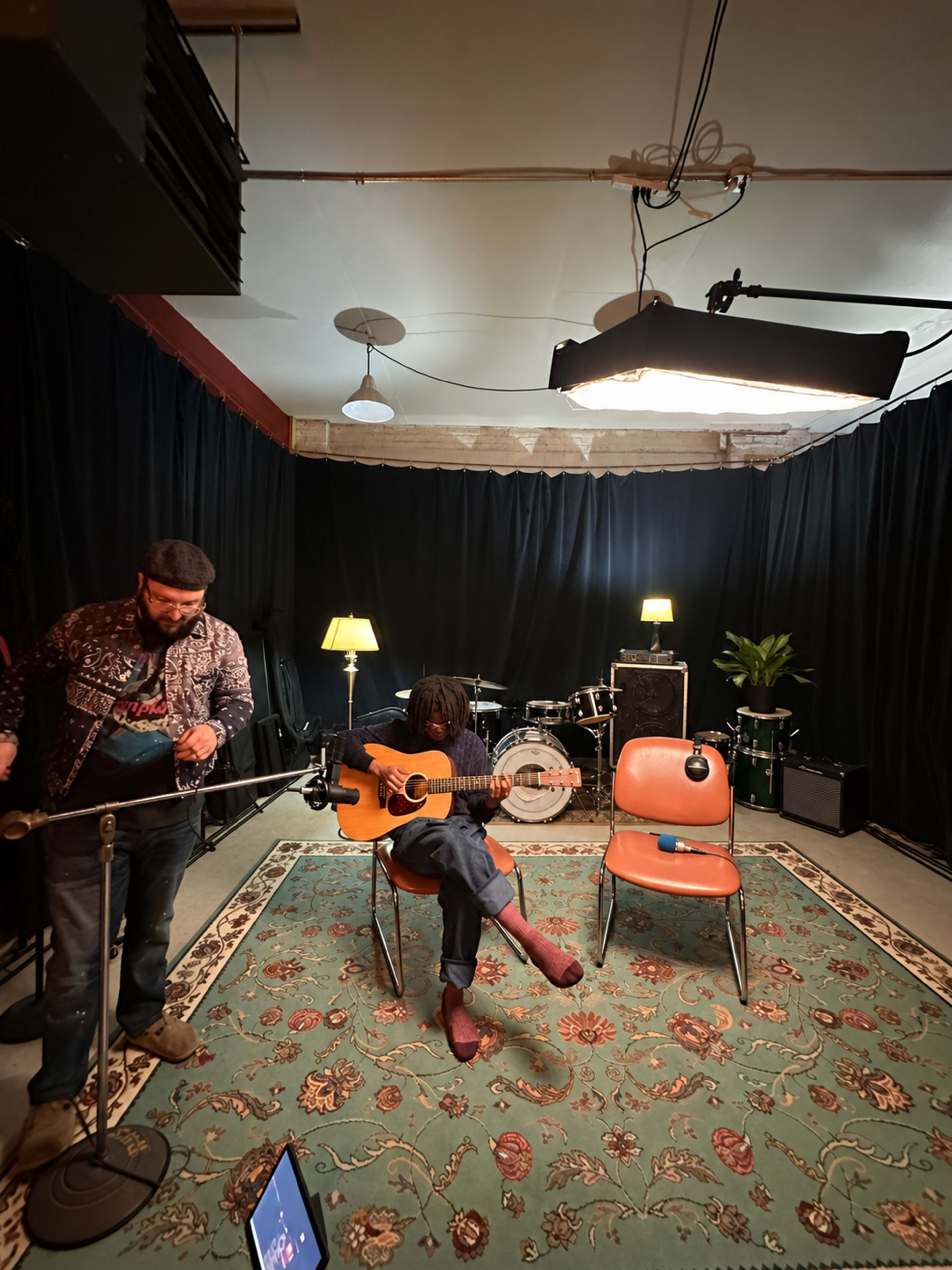 A musician sits on a chair playing an acoustic guitar in a dimly lit room with black curtains, while another person stands nearby with a microphone.