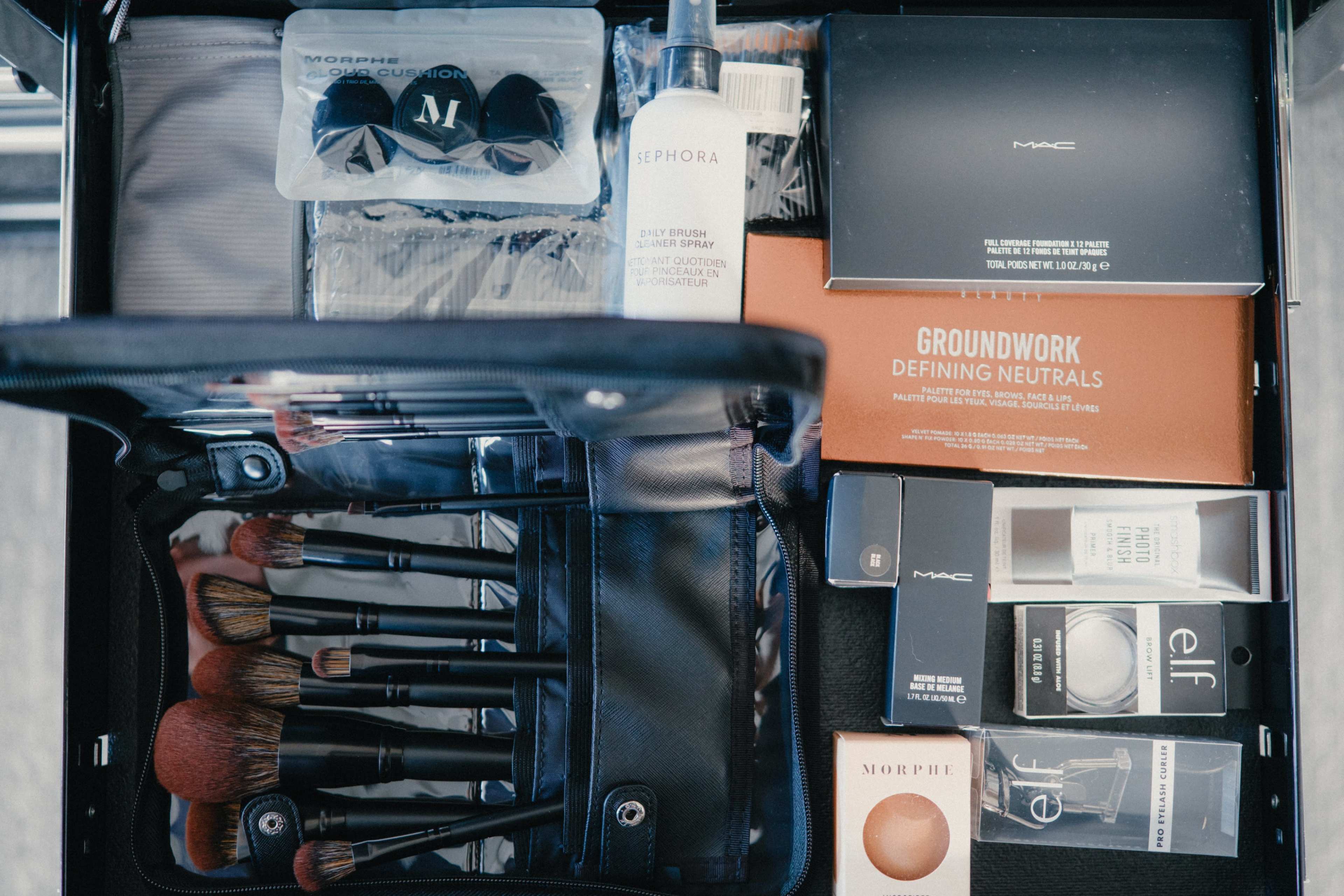 The image shows an open makeup bag containing various beauty products, including brushes, setting sprays, and palettes from brands like MAC, Sephora, and Elf.
