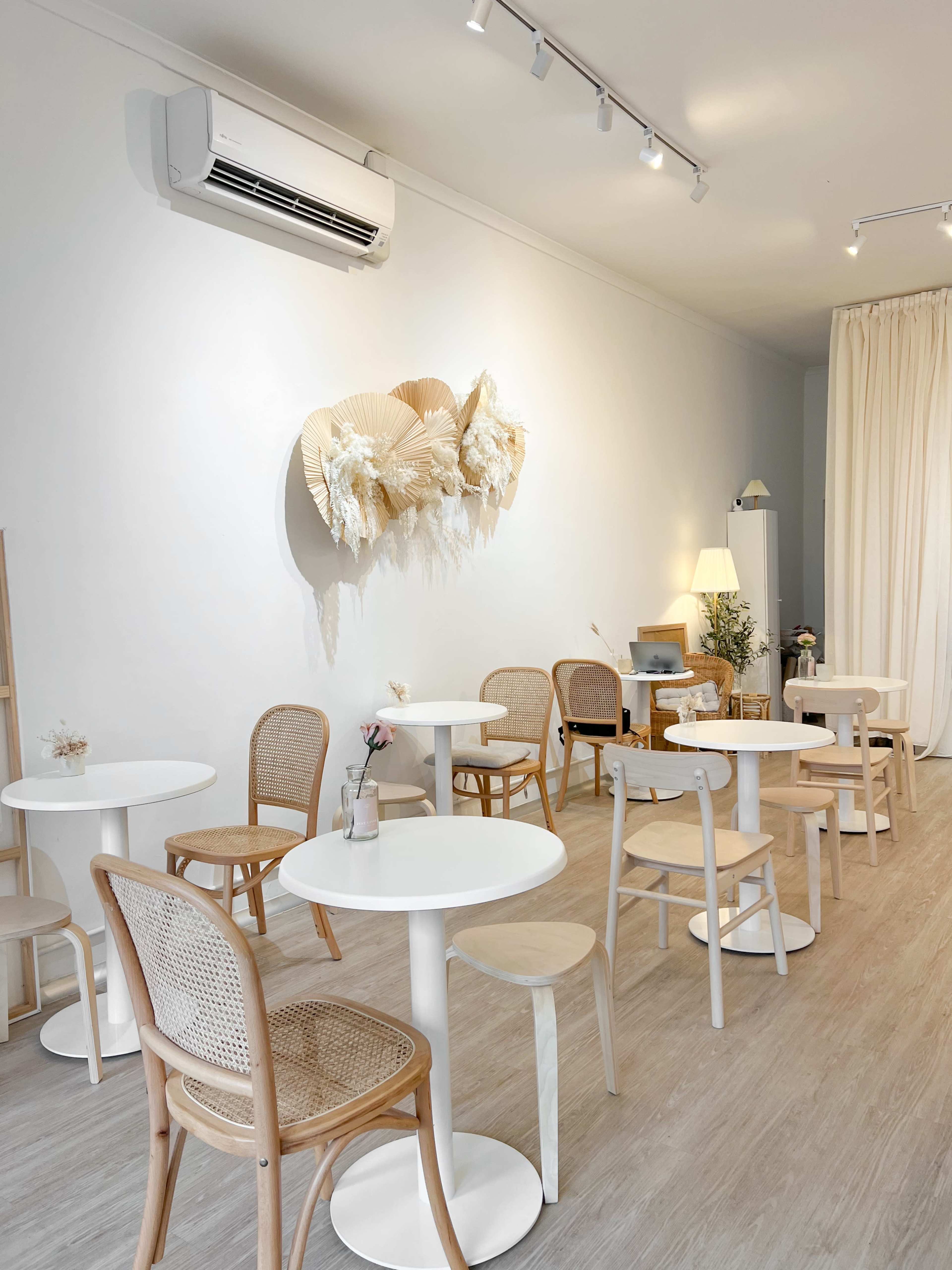 A minimalist café interior with white circular tables and wooden chairs arranged around the space, featuring a decorative wall piece made of dried plants.