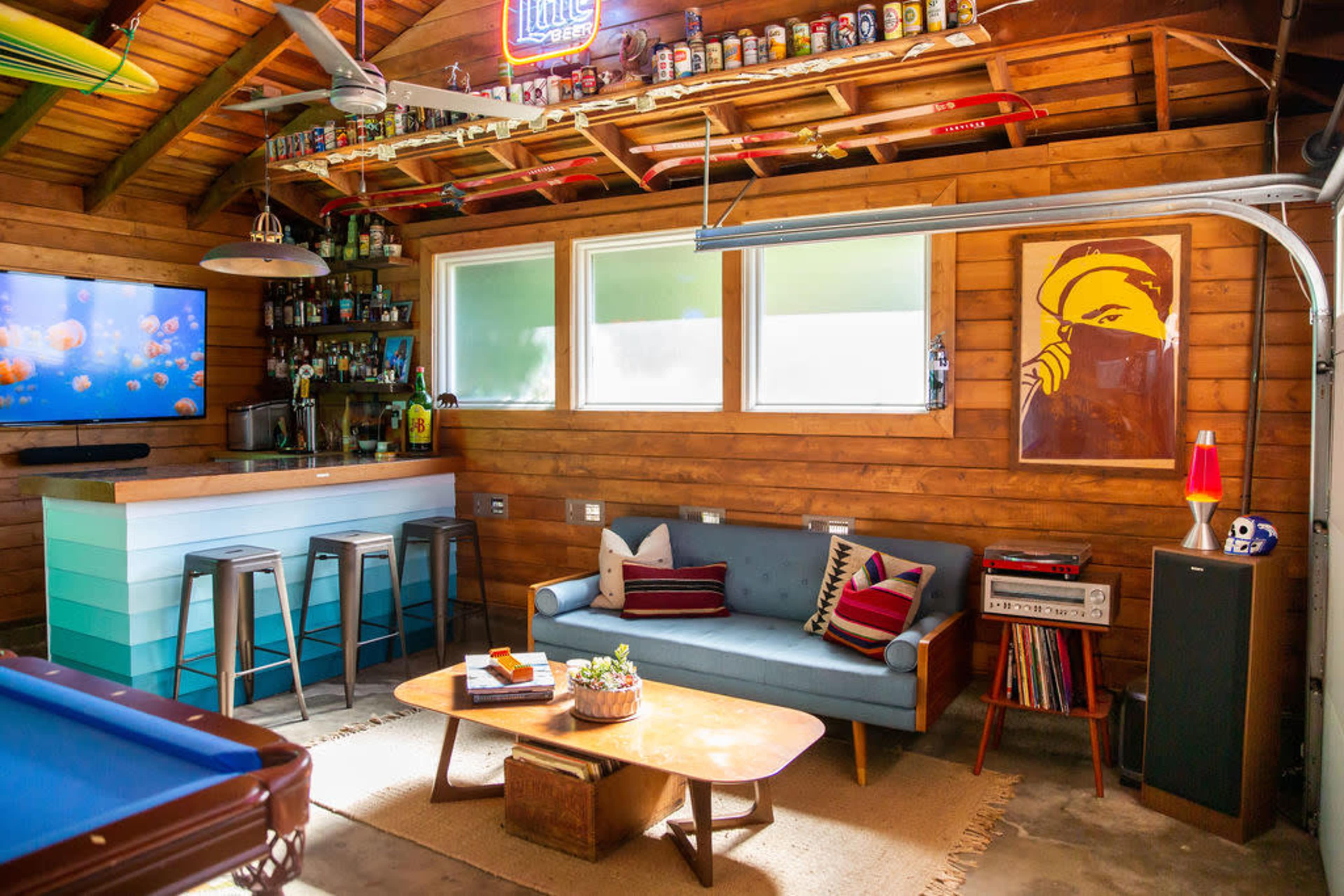 A cozy, rustic room featuring a bar area, a comfortable couch with decorative pillows, a coffee table, a pool table, and a wall decorated with artwork and shelves filled with various items.
