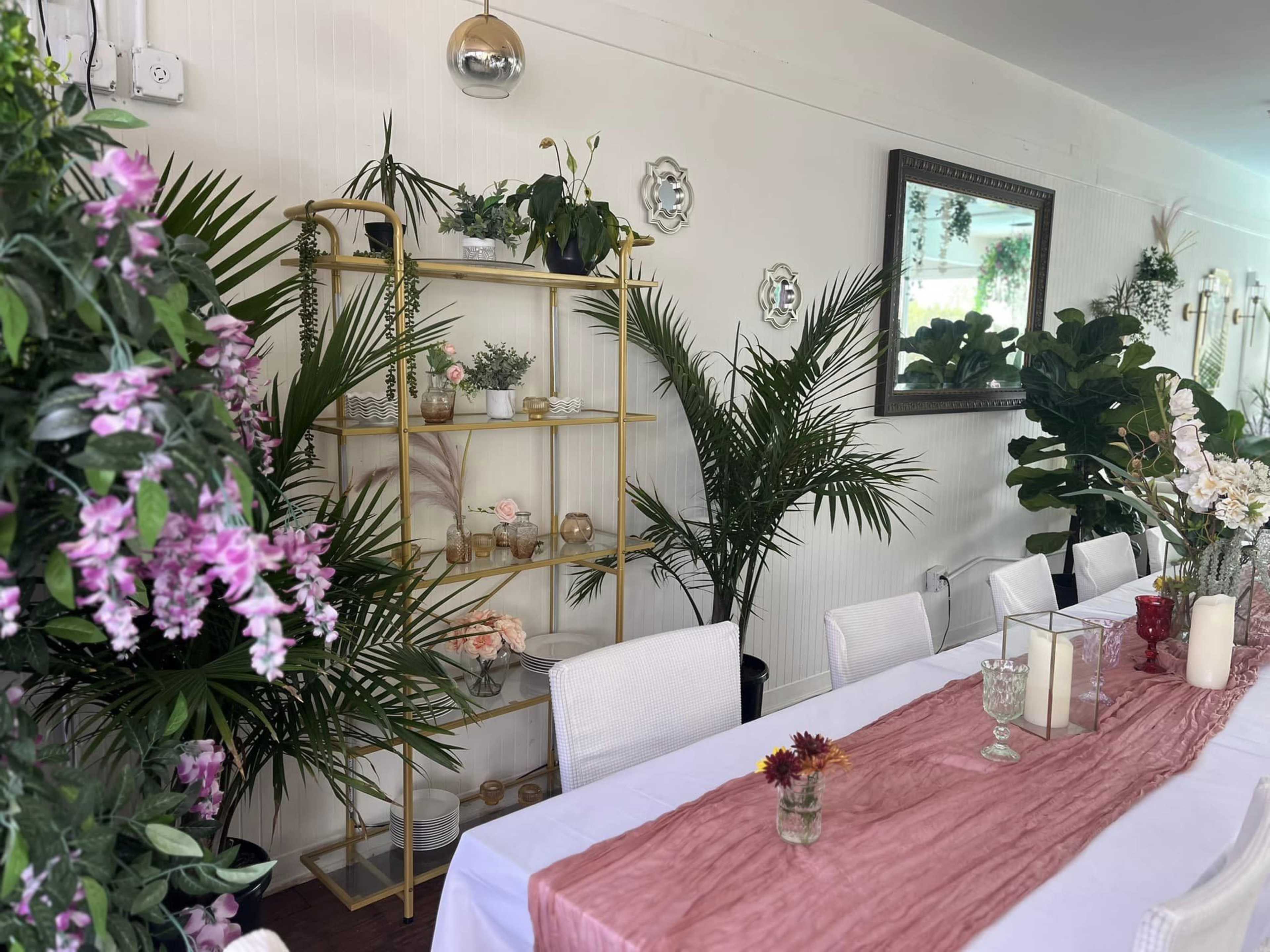 A long dining table is set with a pink runner and surrounded by white chairs, with shelves displaying plants and decorative items in a room adorned with greenery and floral arrangements.