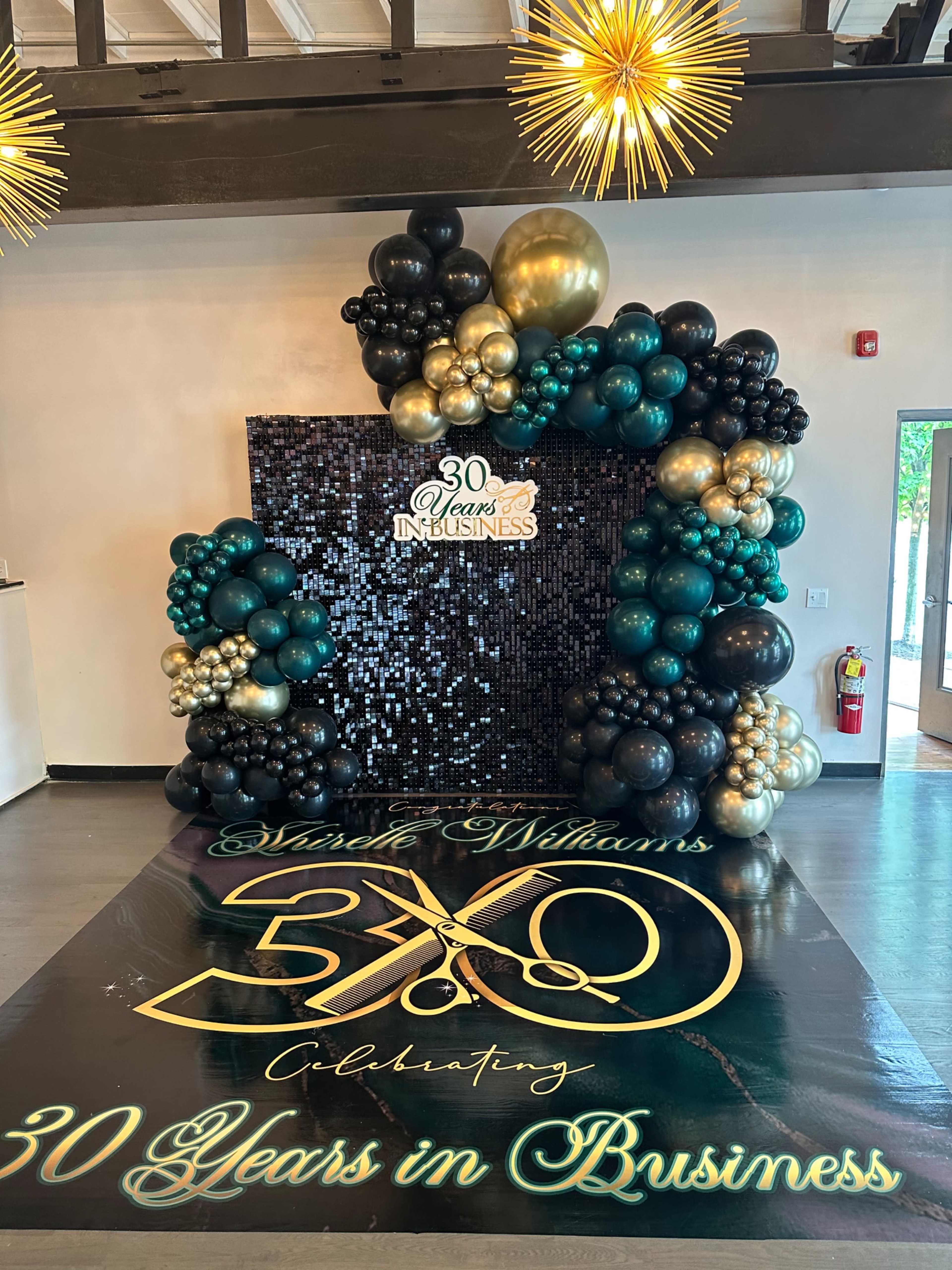 A decorative backdrop for a celebration, with a large arrangement of black, gold, and teal balloons and a banner stating "30 Years in Business" alongside the name "Marcia Williams."