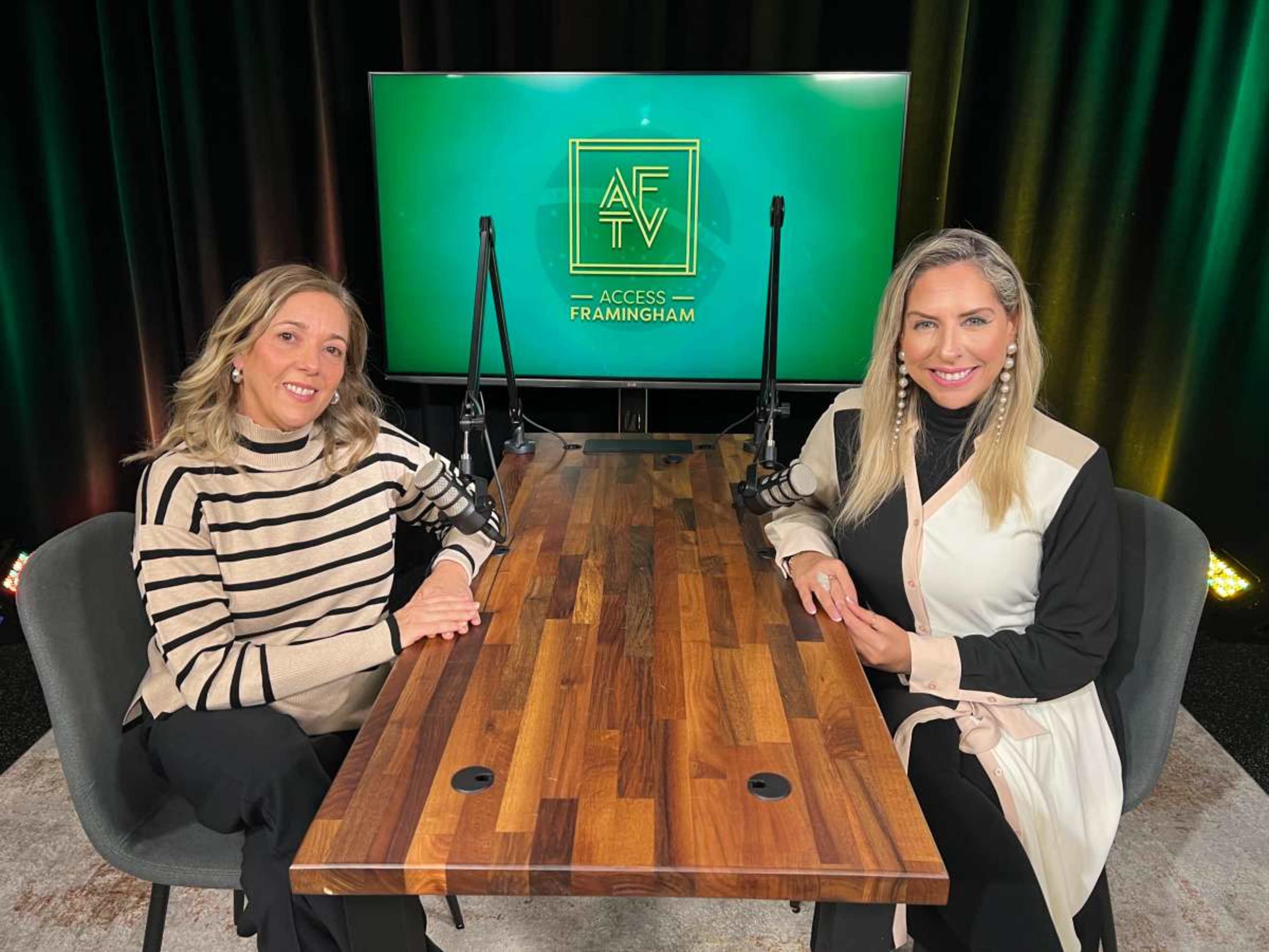 Two women are sitting at a wooden table in a modern studio with a green backdrop featuring the logo for "Access Framingham."