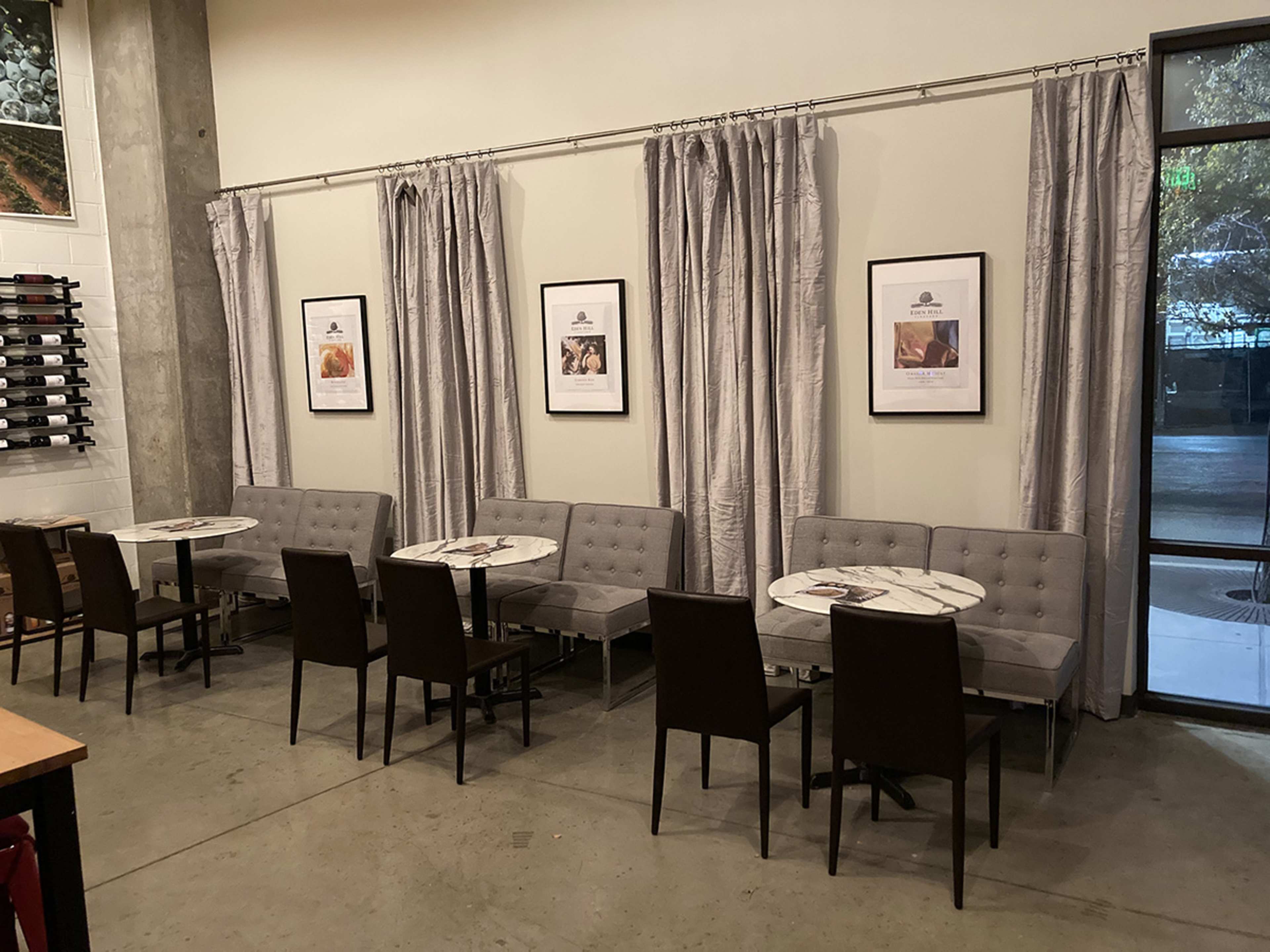 A restaurant interior with several gray benches and tables arranged against a wall, accompanied by framed pictures and floor-to-ceiling curtains.
