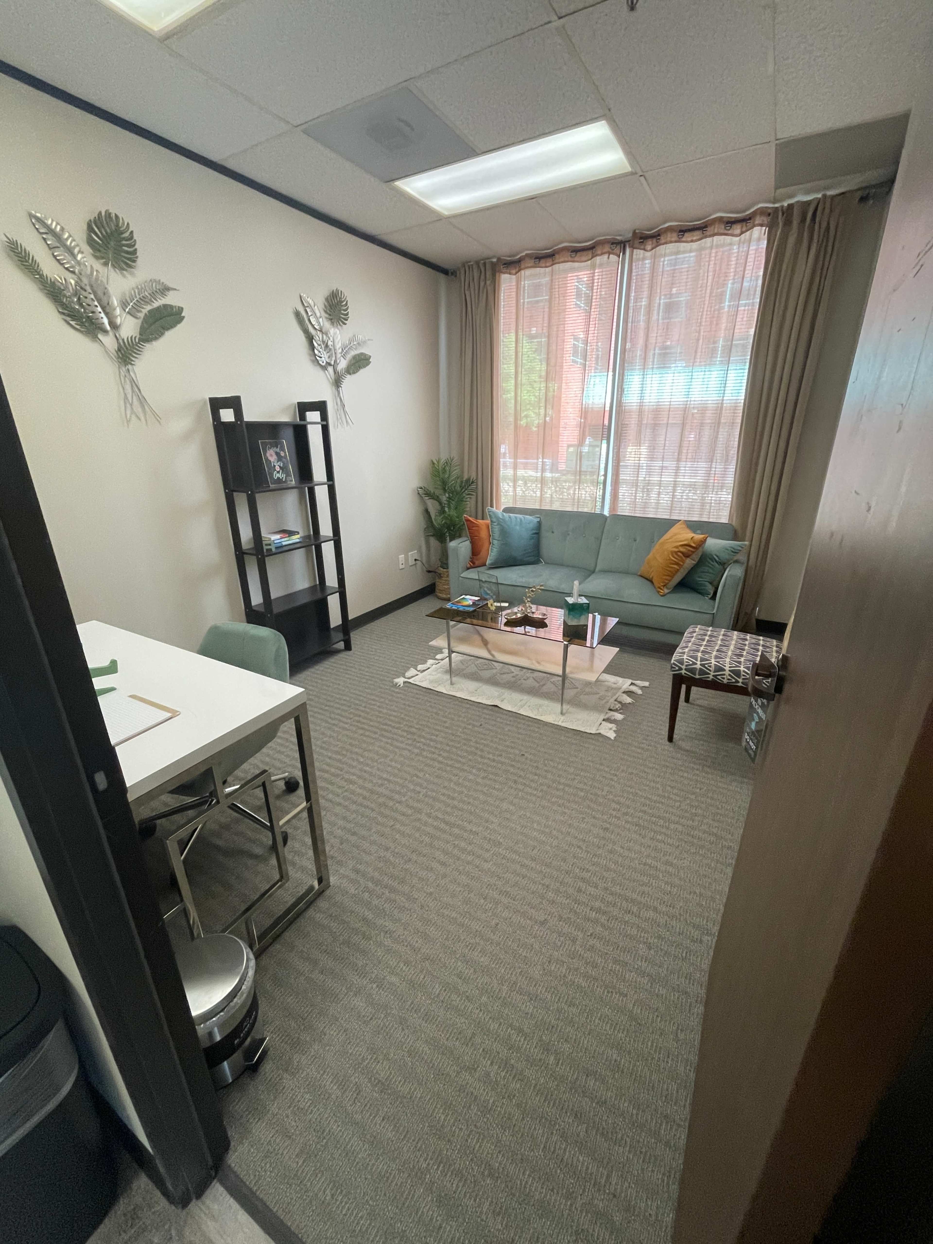 A professional office space with a green couch, a coffee table, a small bookshelf, and a desk with chairs, bathed in natural light from a window.