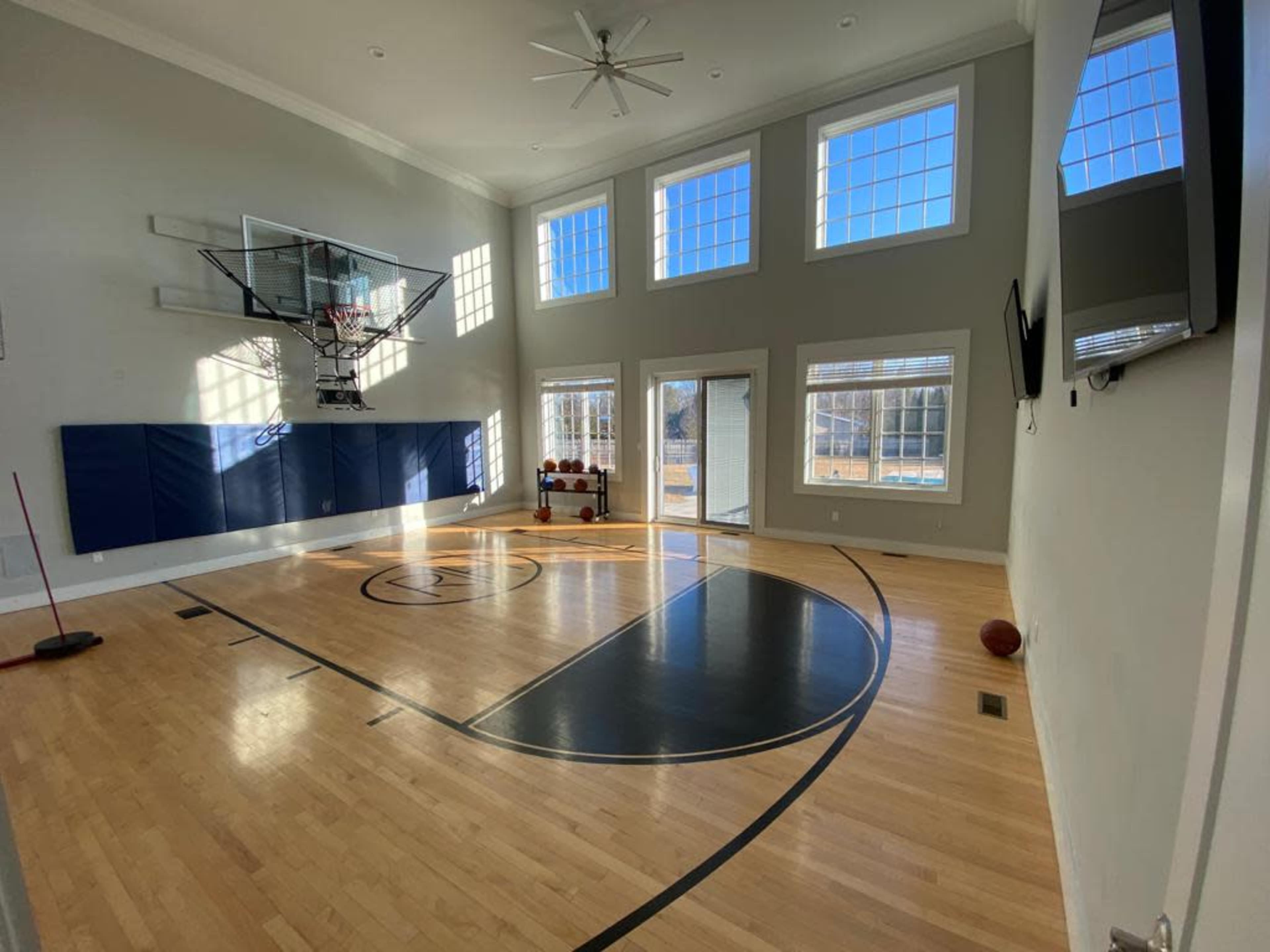 The image shows an indoor basketball court with large windows, a hoop mounted on the wall, and a hardwood floor featuring a center court design.