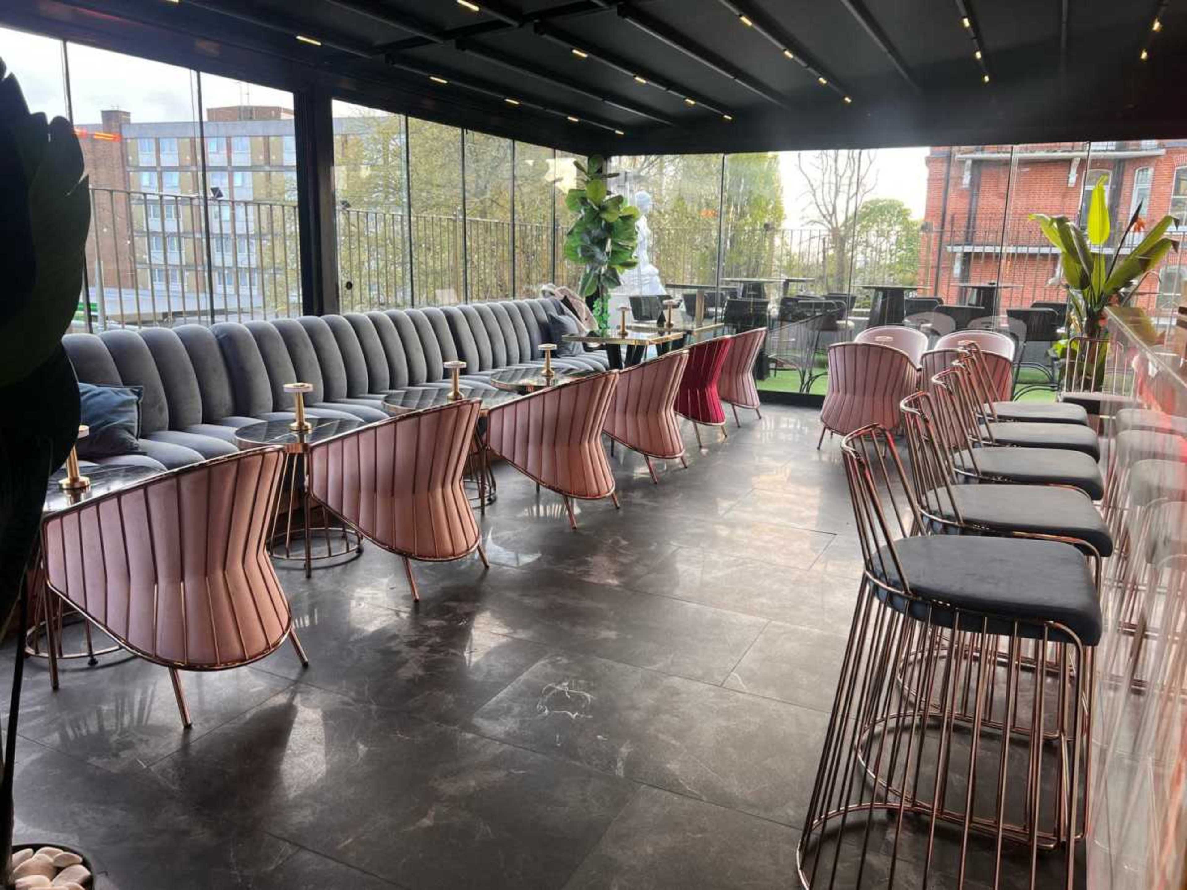 The image shows a modern indoor seating area with plush seating and stylish tables, surrounded by large windows and greenery.
