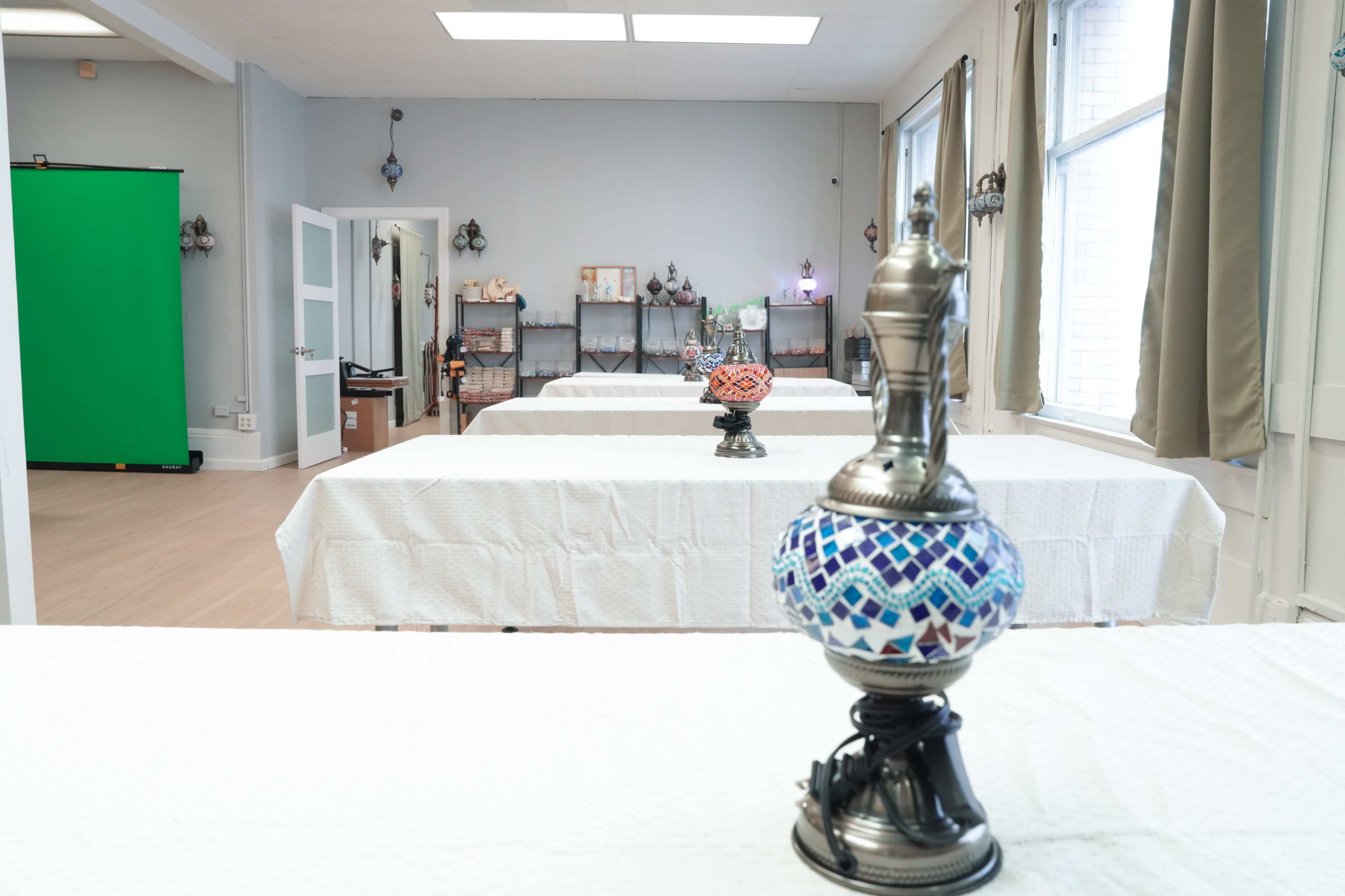 The image shows a spacious room with multiple tables covered in white tablecloths, each decorated with a small, ornate lamp, and a green screen in the background.