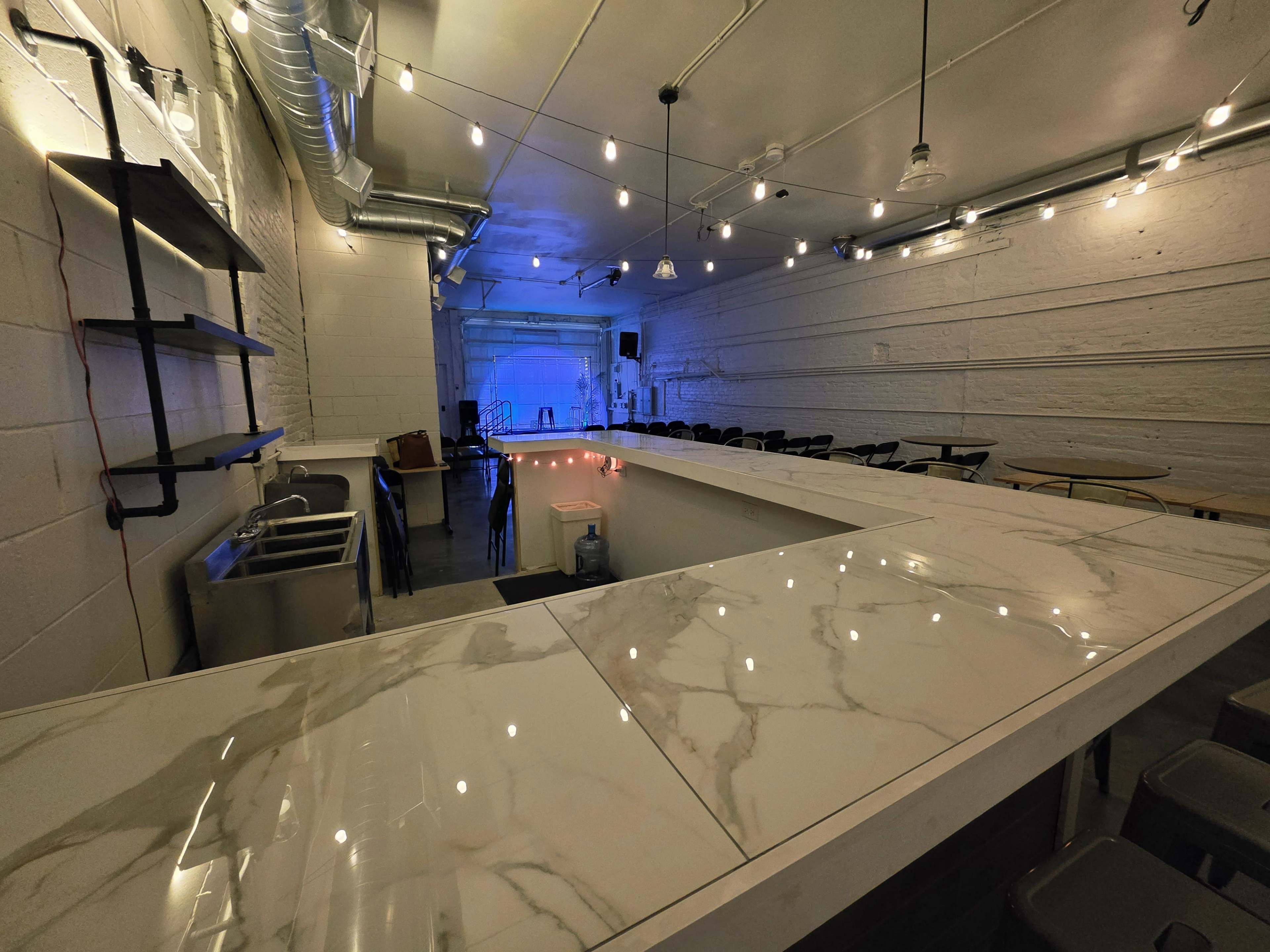 The image shows a spacious, modern bar area with a marble countertop, metal shelves, and a dimly lit performance space in the background.