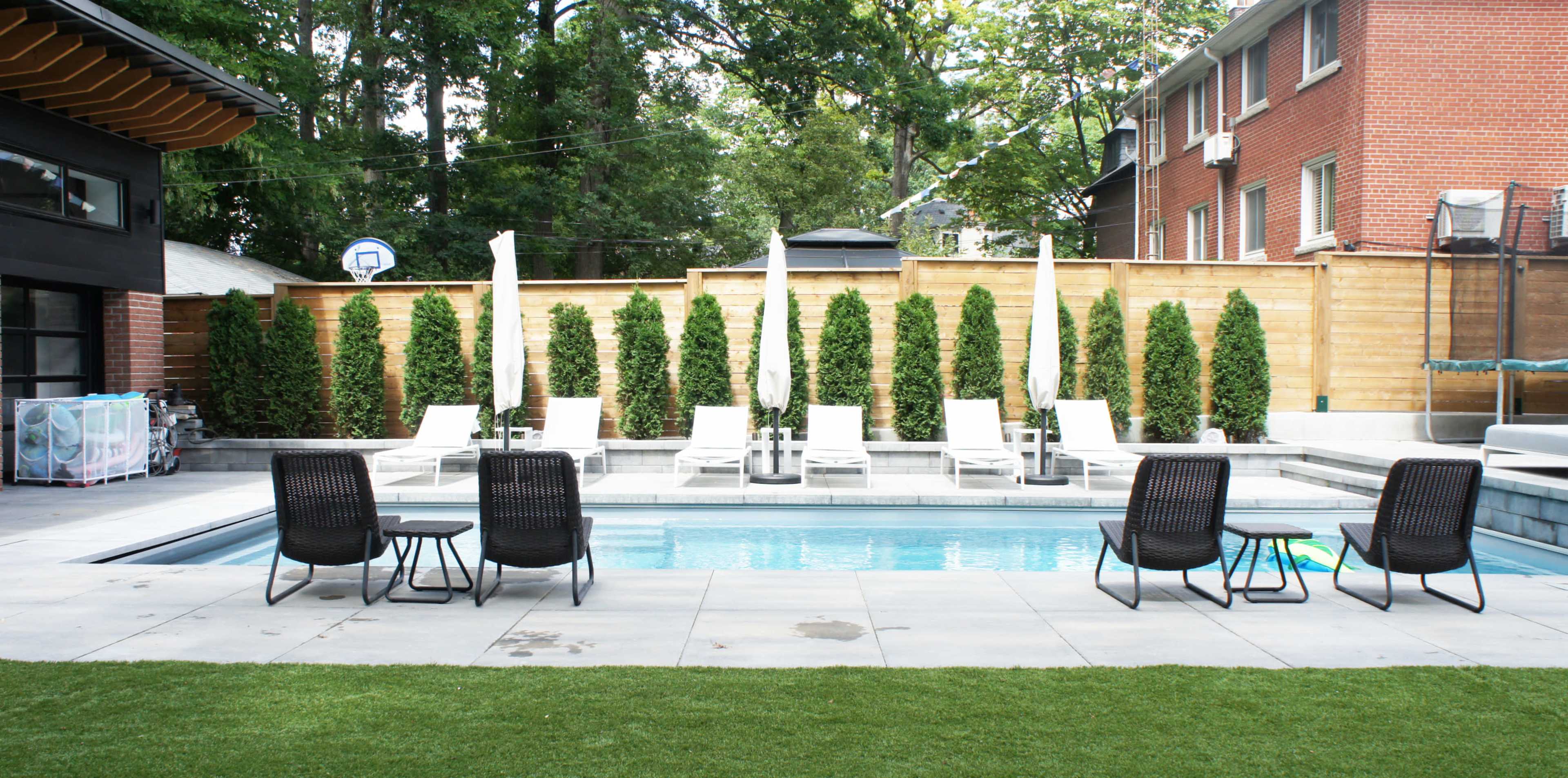 A modern backyard features a rectangular swimming pool surrounded by lounge chairs, with tall greenery and a wooden fence in the background.
