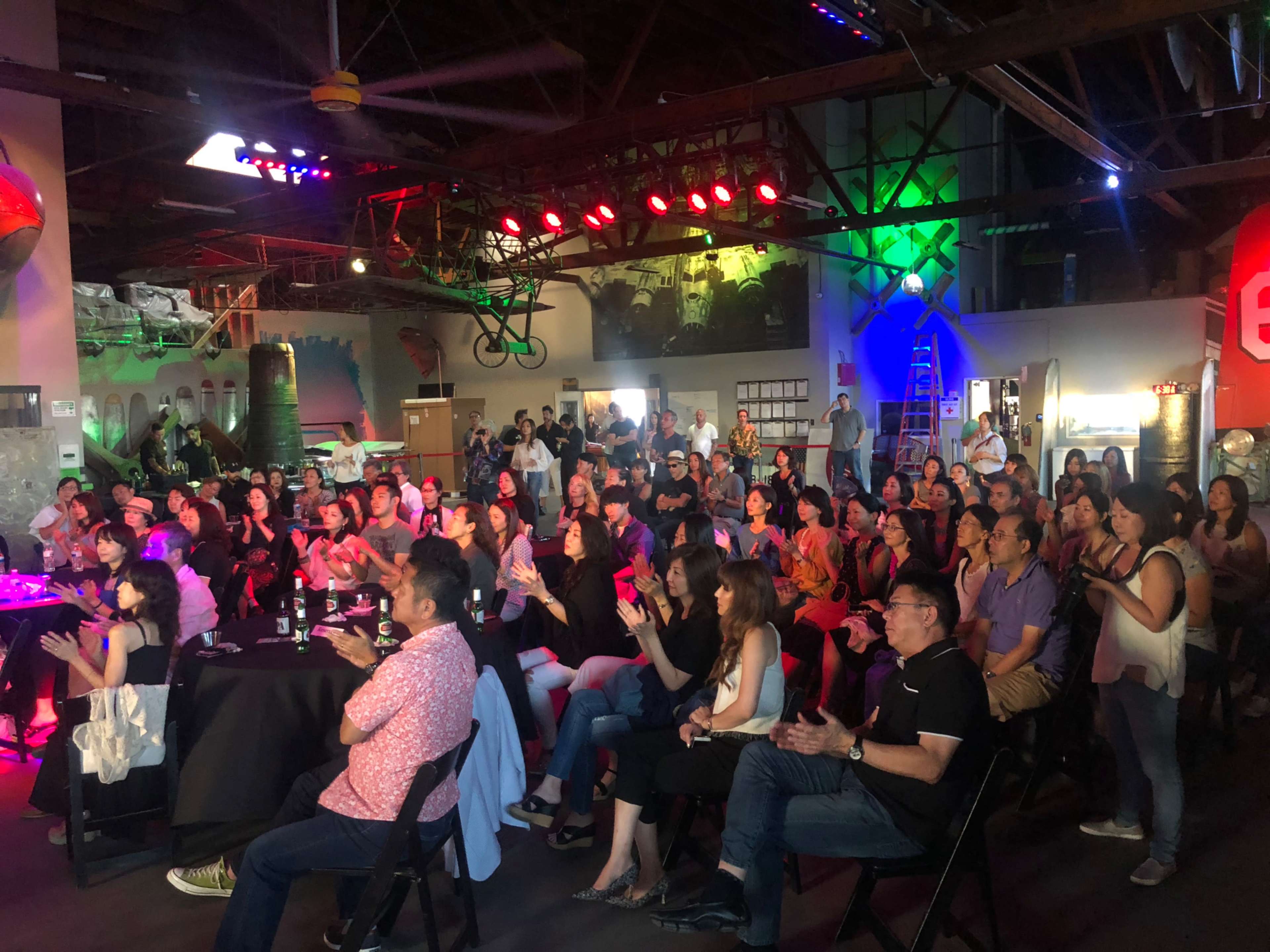 A large audience is seated at tables in a dimly lit venue, with colorful lights illuminating the space and a stage in view.