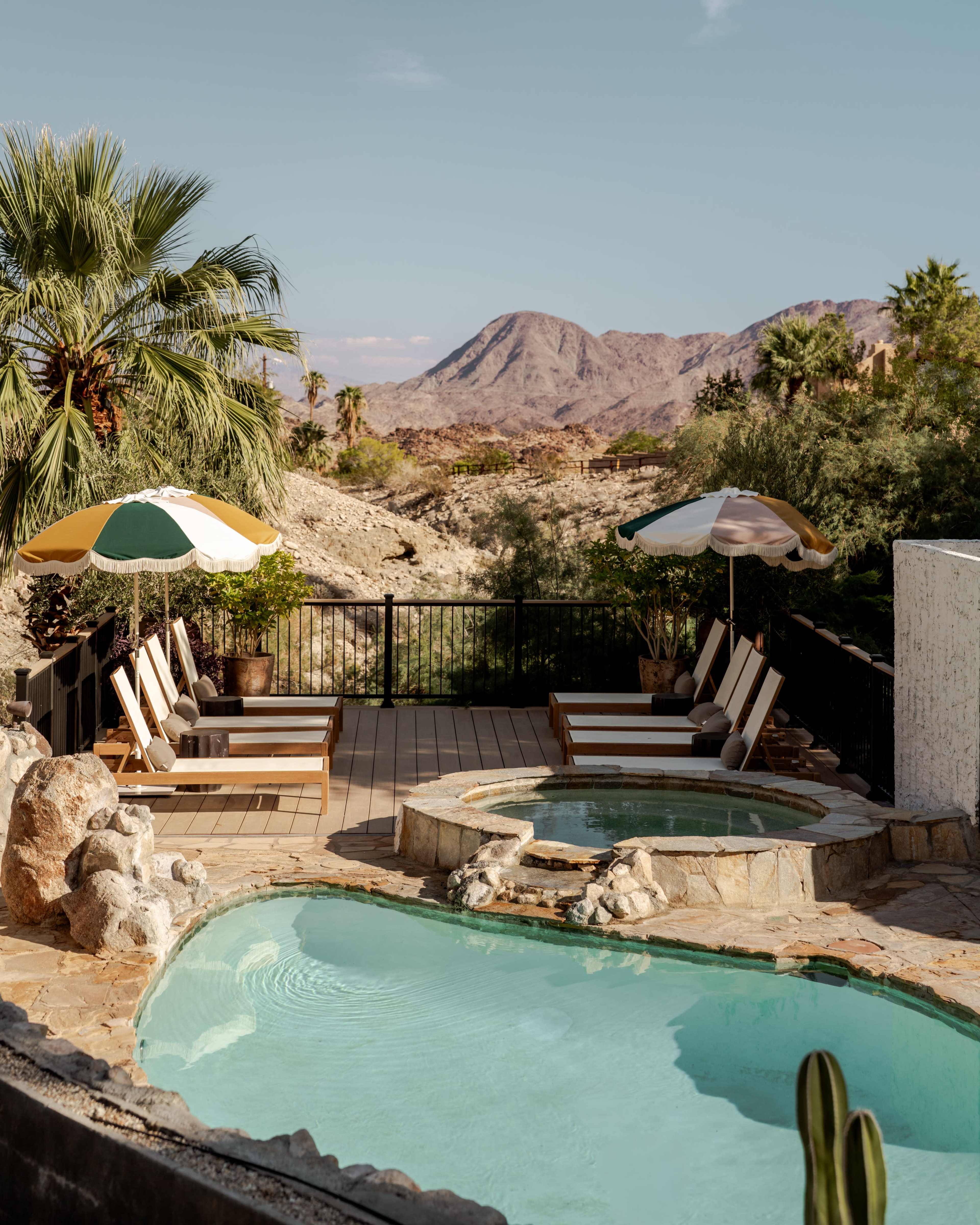 The image shows a peaceful outdoor space featuring a swimming pool and a hot tub, surrounded by lounge chairs and palm trees, with mountains visible in the background.