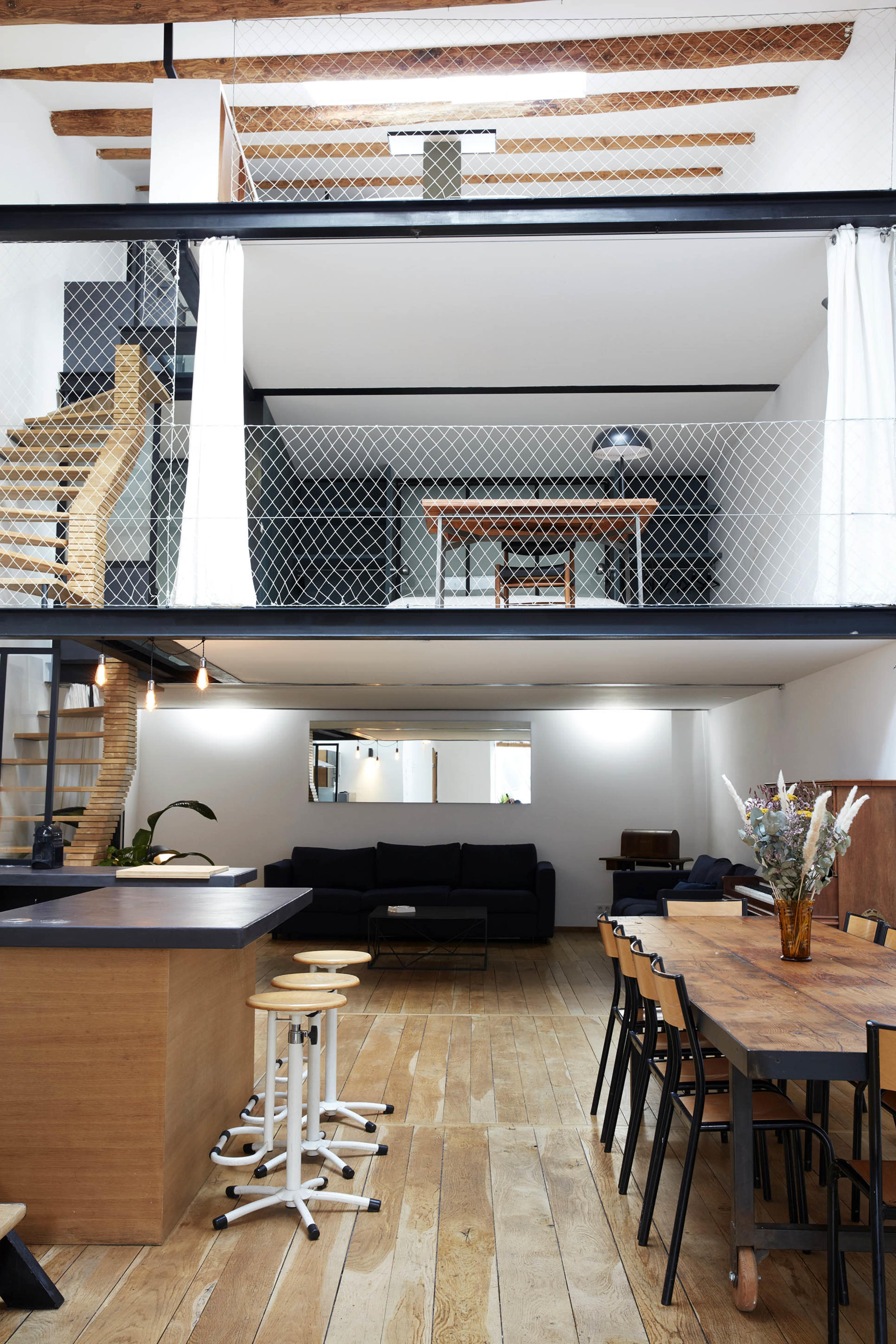 The image shows an open-concept living space featuring a kitchen with a wooden island, a dining area with black chairs, and a lofted area with a wooden table overlooking the main floor.