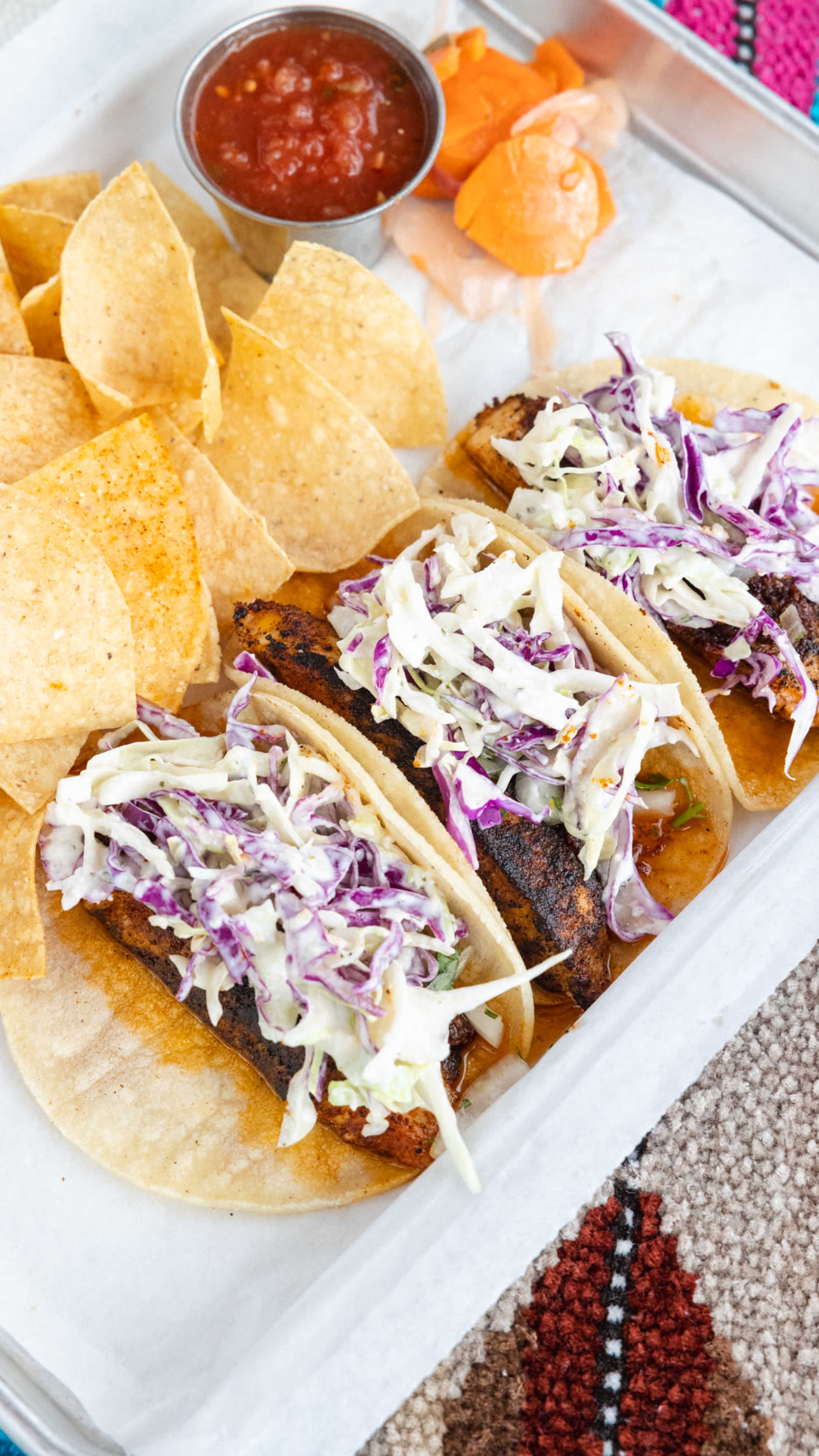 The image shows two tacos filled with grilled fish and topped with cabbage slaw, accompanied by tortilla chips, salsa, and a side of orange slices on a tray.