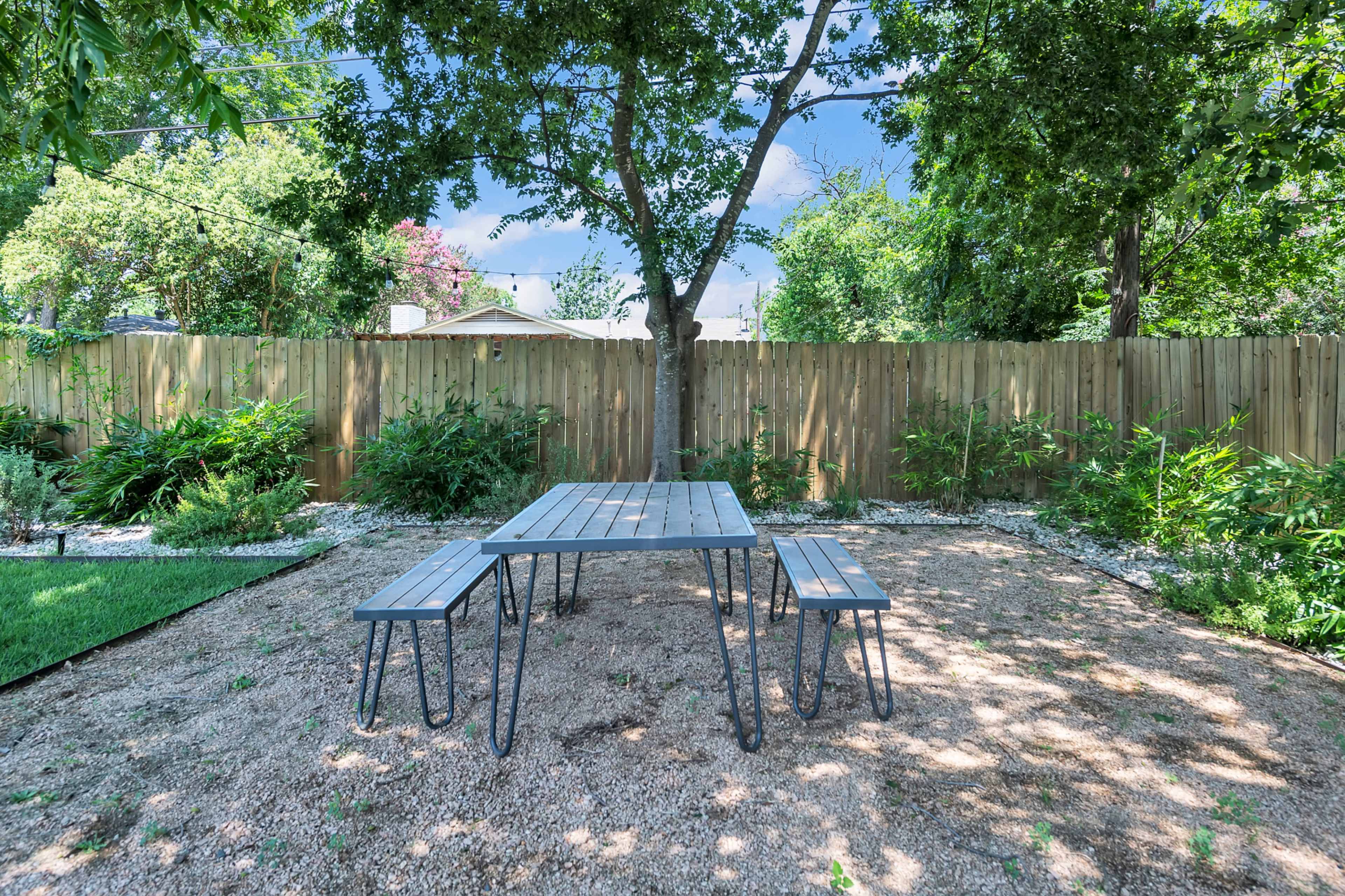 A metal table with two benches is positioned on gravel in a fenced backyard surrounded by greenery.