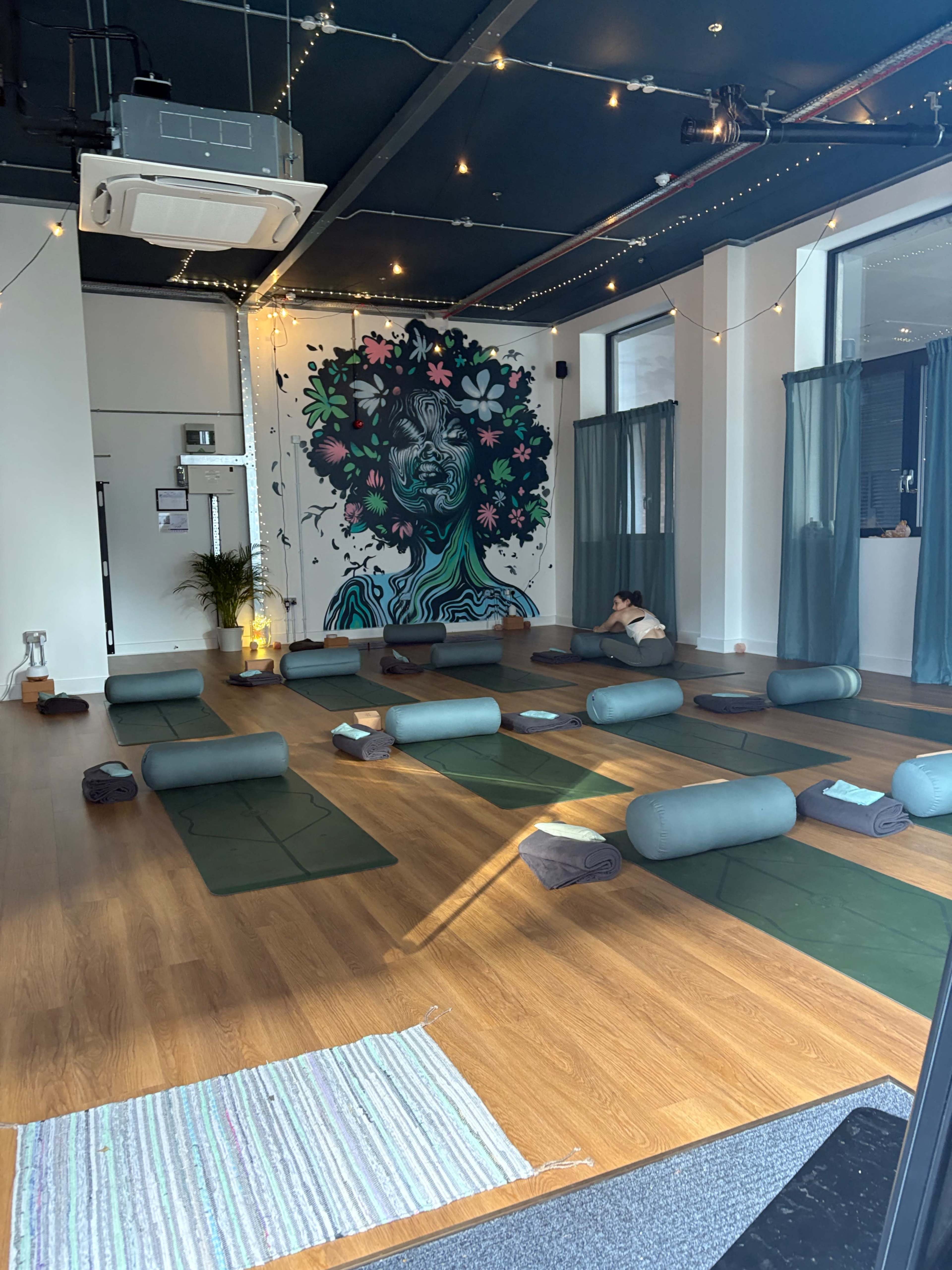 The image shows a spacious yoga studio featuring green mats arranged on a wooden floor, a mural of a woman with floral designs on a prominent wall, and soft lighting.