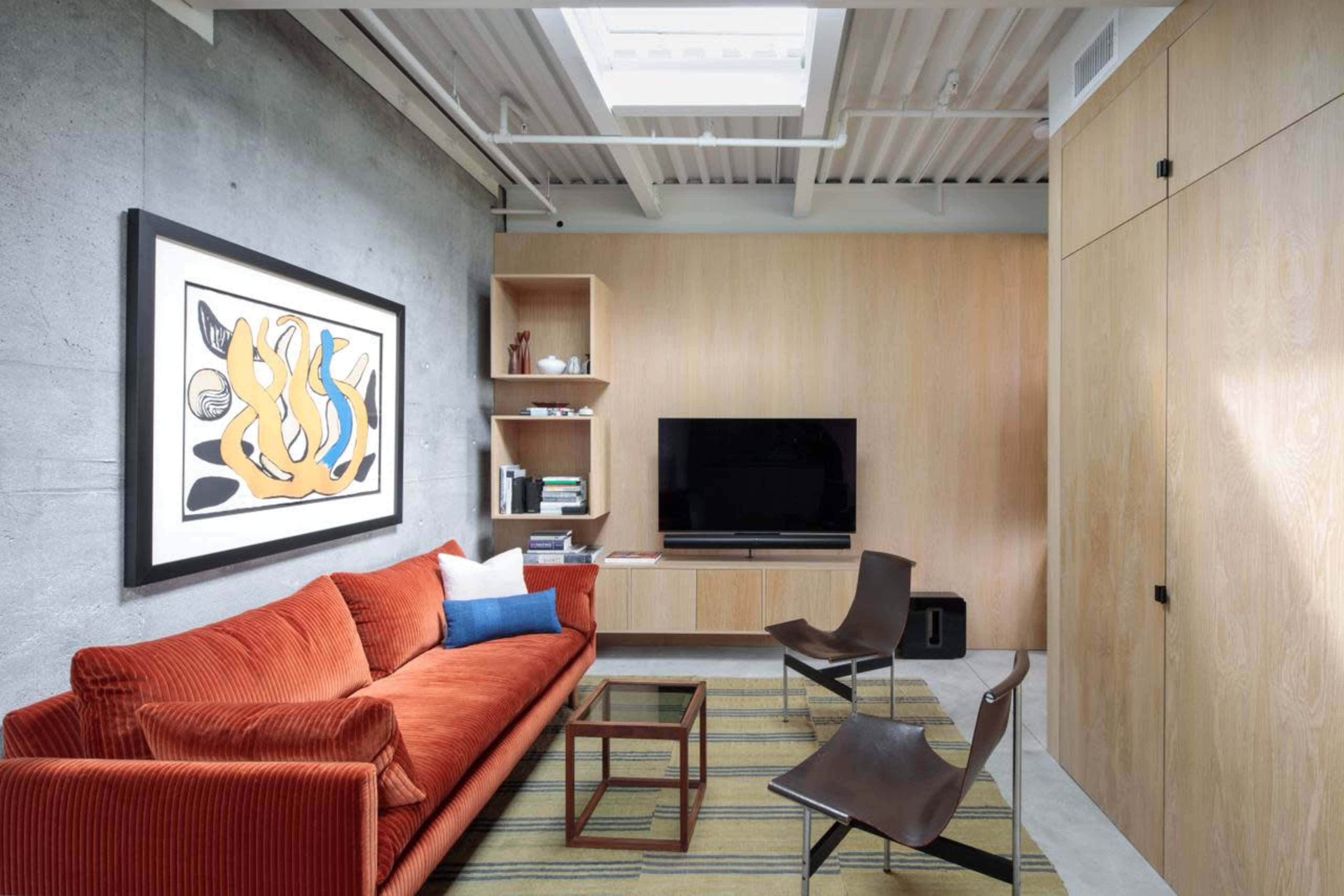 The image shows a modern living room with a red sofa, a television mounted on a wooden wall unit, and a skylight overhead.