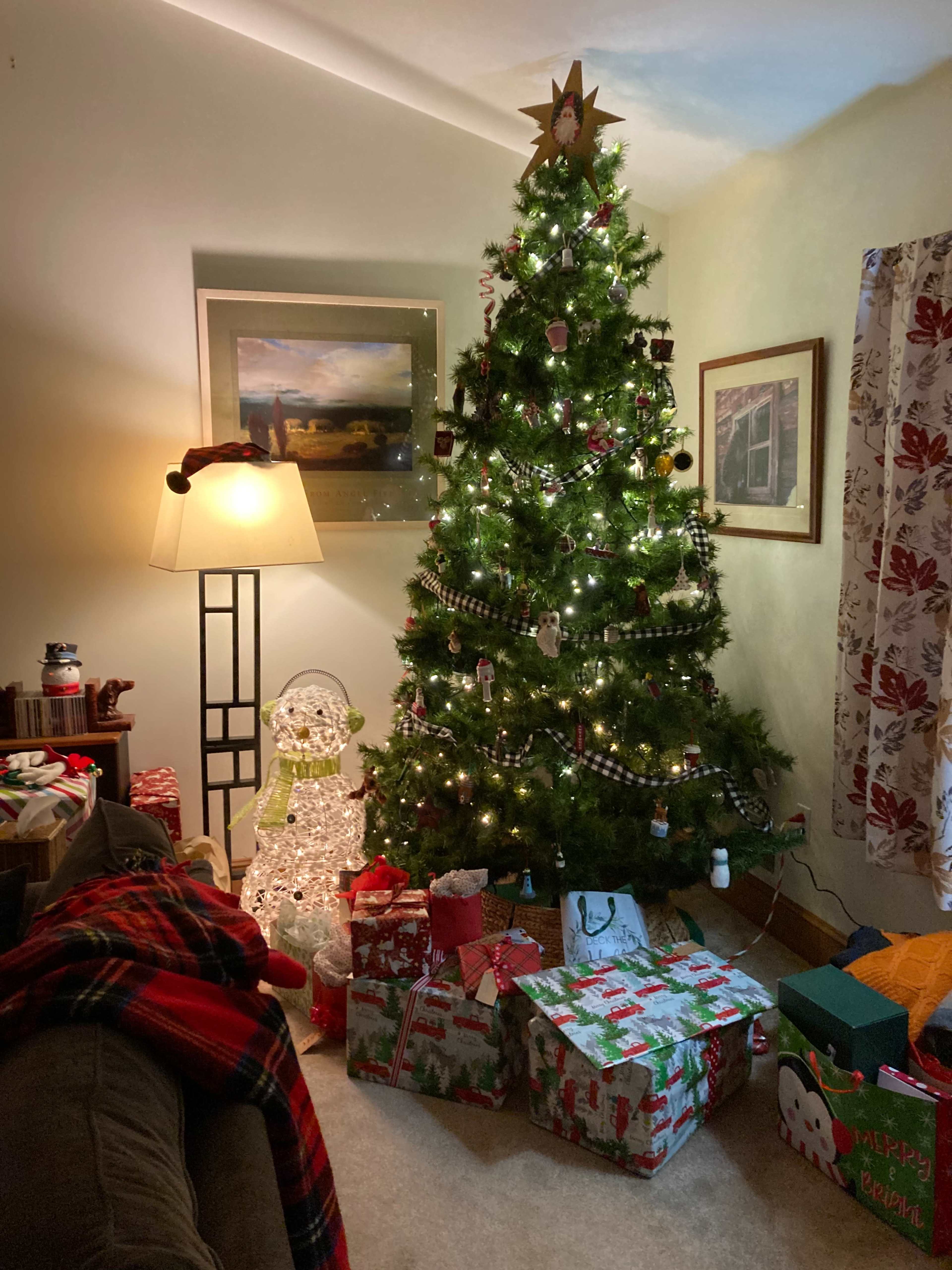 A decorated Christmas tree stands in a cozy living room surrounded by wrapped gifts and a lighted snowman.