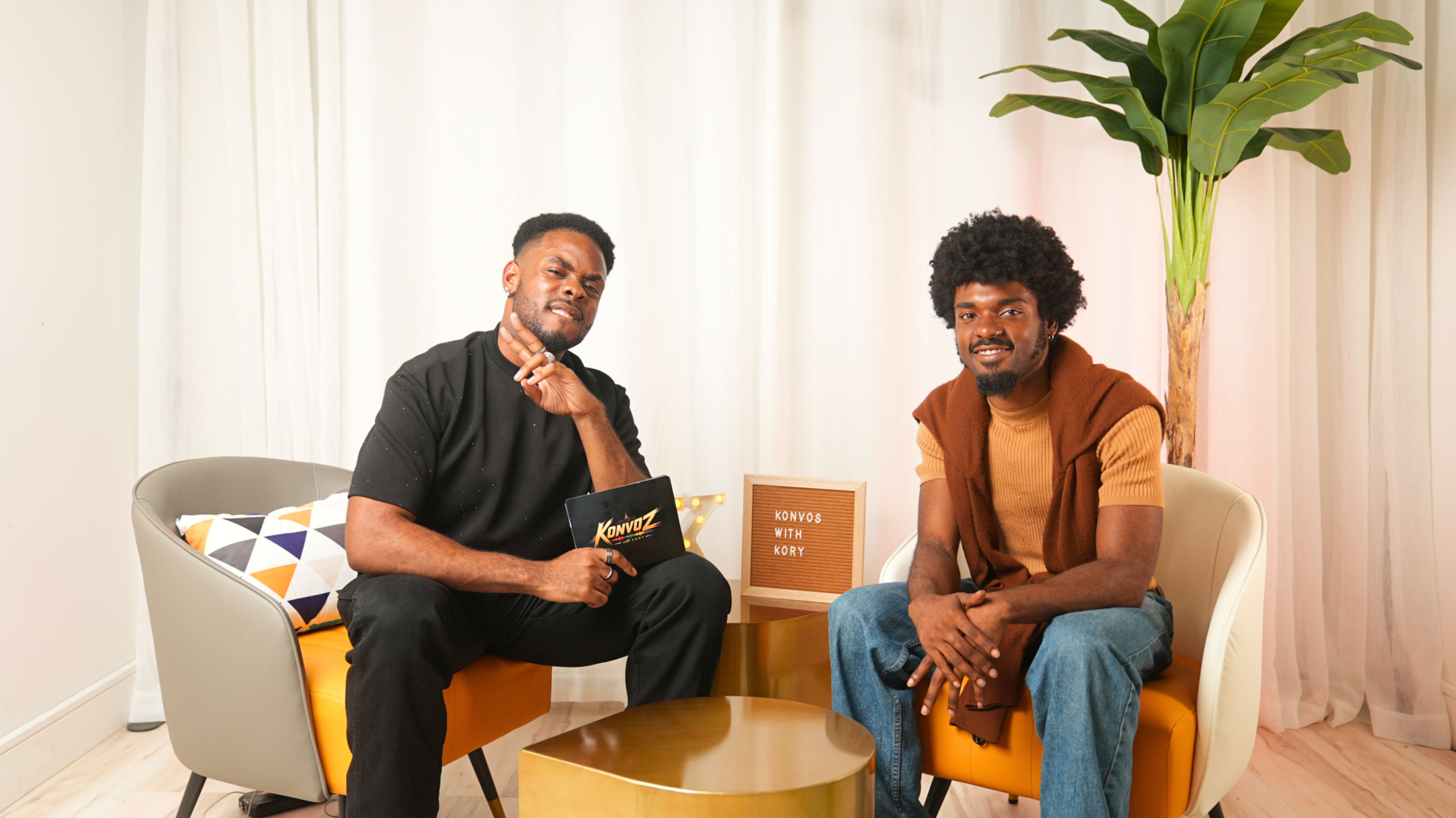 Two men sit in modern chairs in a well-lit room, one holding a sign while the other poses playfully with a finger on his lips.