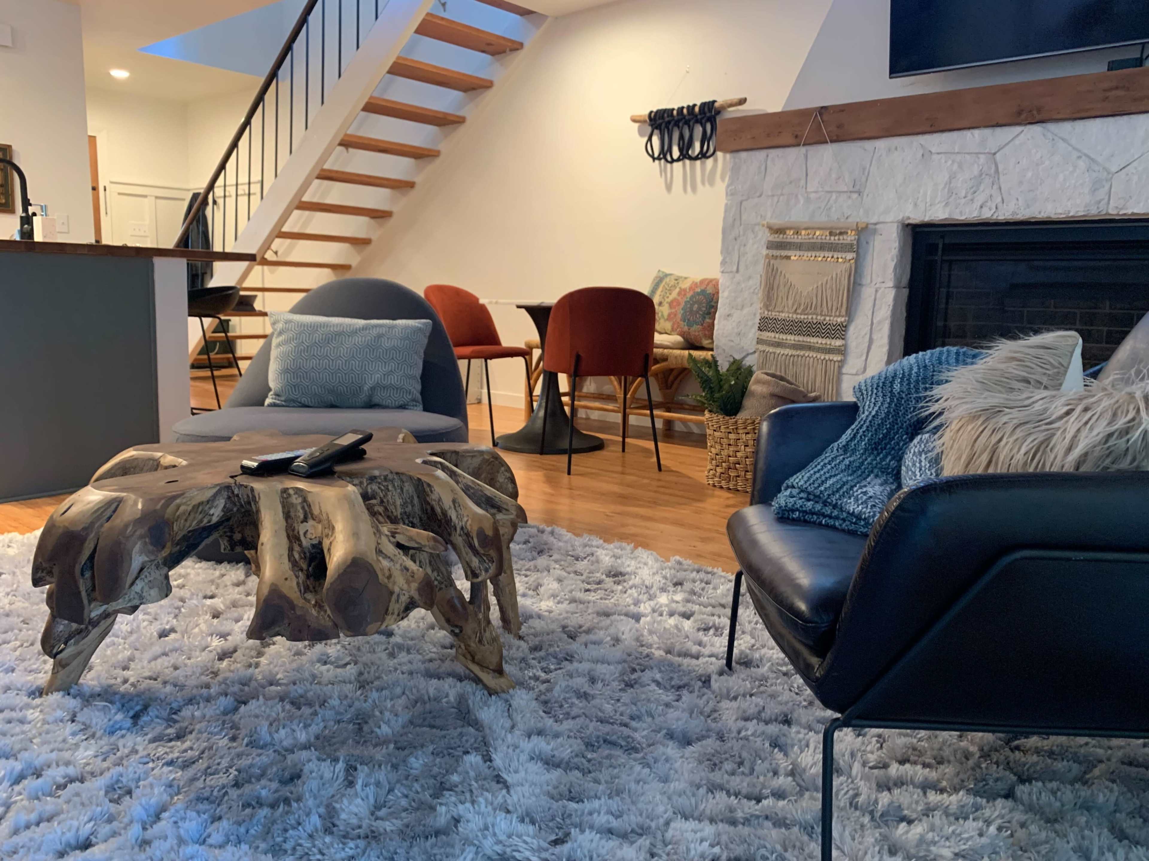 A modern living space featuring a wooden coffee table, two armchairs, a faux fur rug, and a staircase in the background.