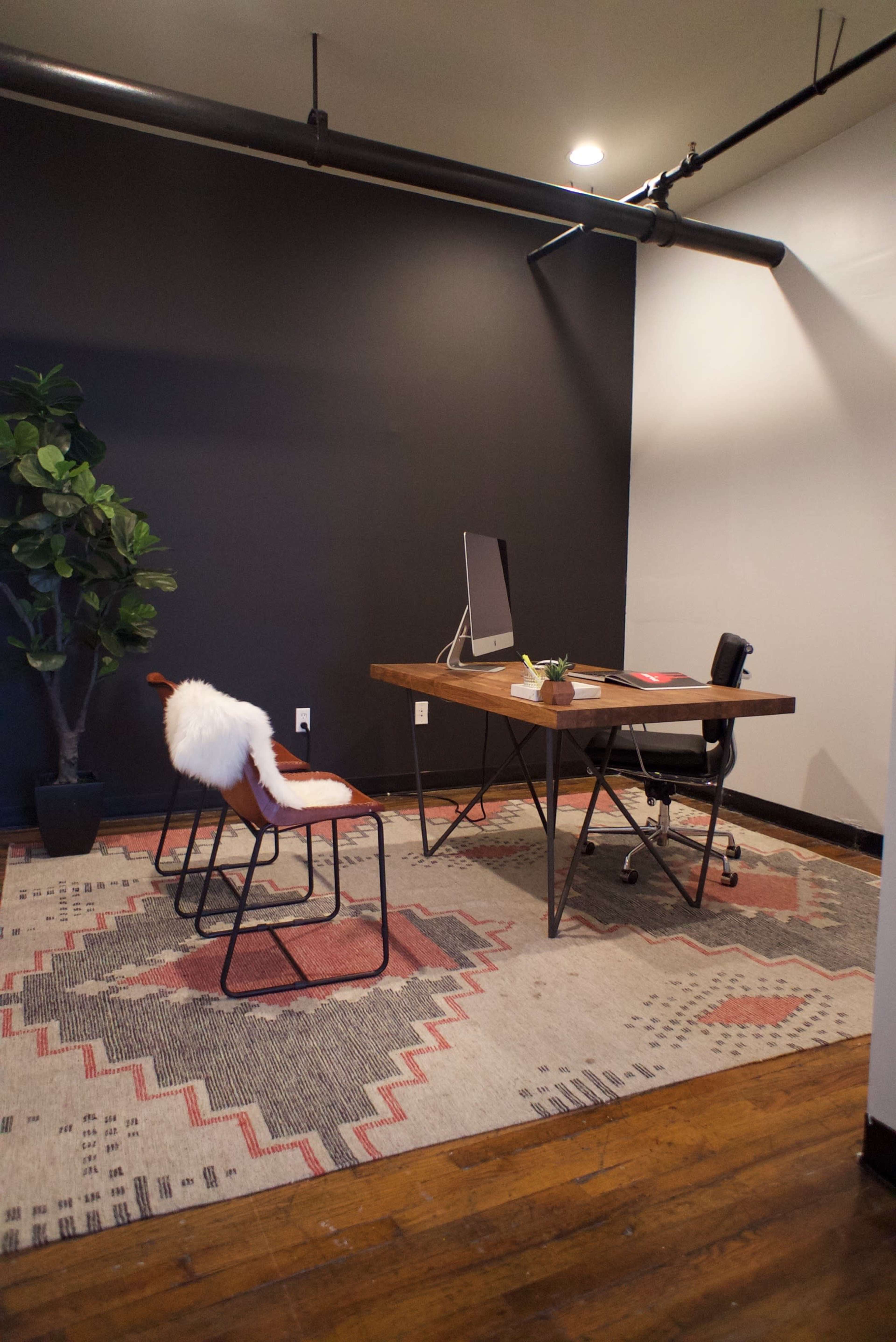 The image shows a modern office space featuring a wooden desk, a computer monitor, a black chair, and a decorative plant, with a patterned rug on the wooden floor.
