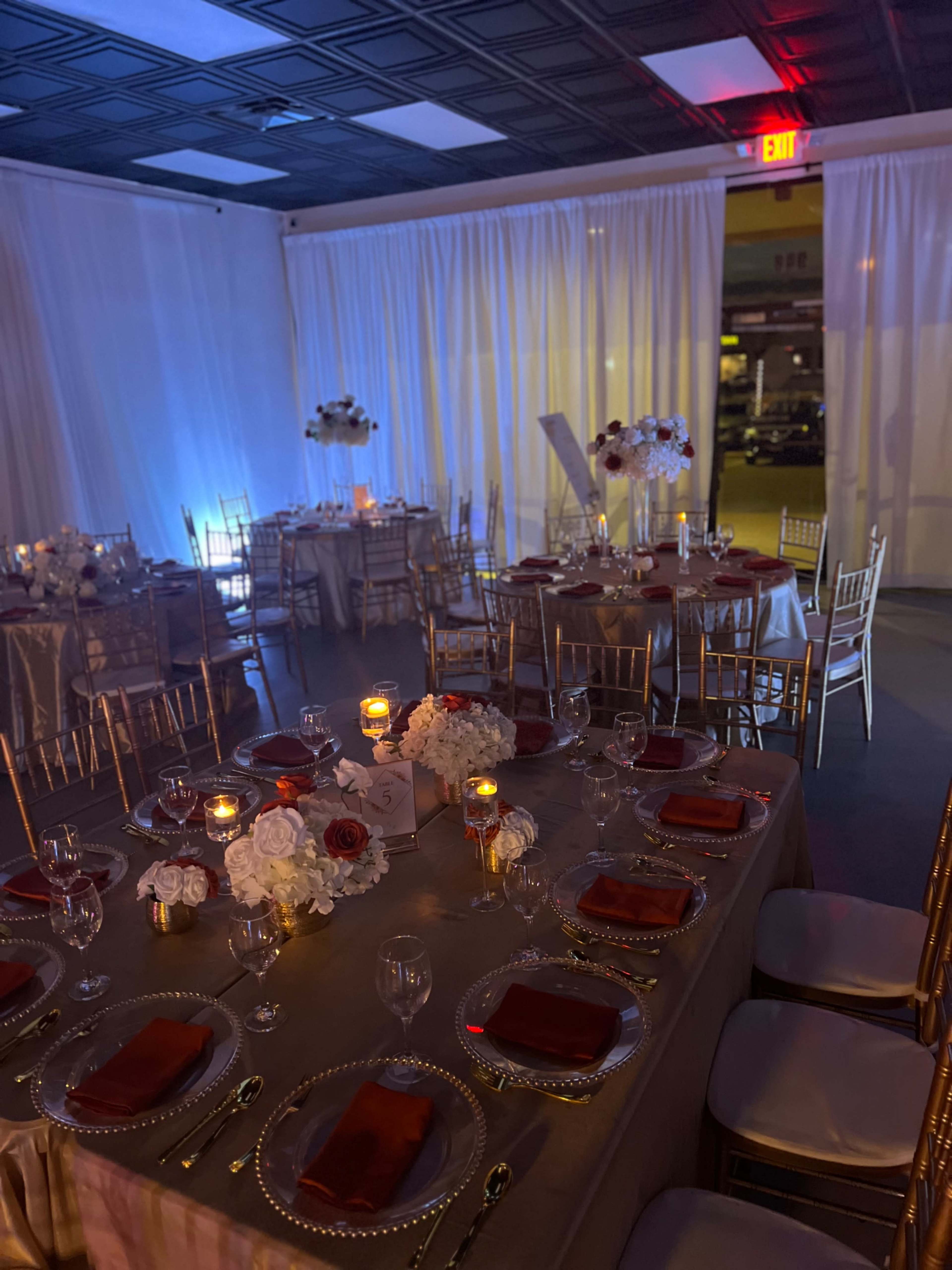 A banquet setup with elegantly arranged tables featuring gold chairs, white tablecloths, and centerpieces of flowers and candles, all under soft lighting.