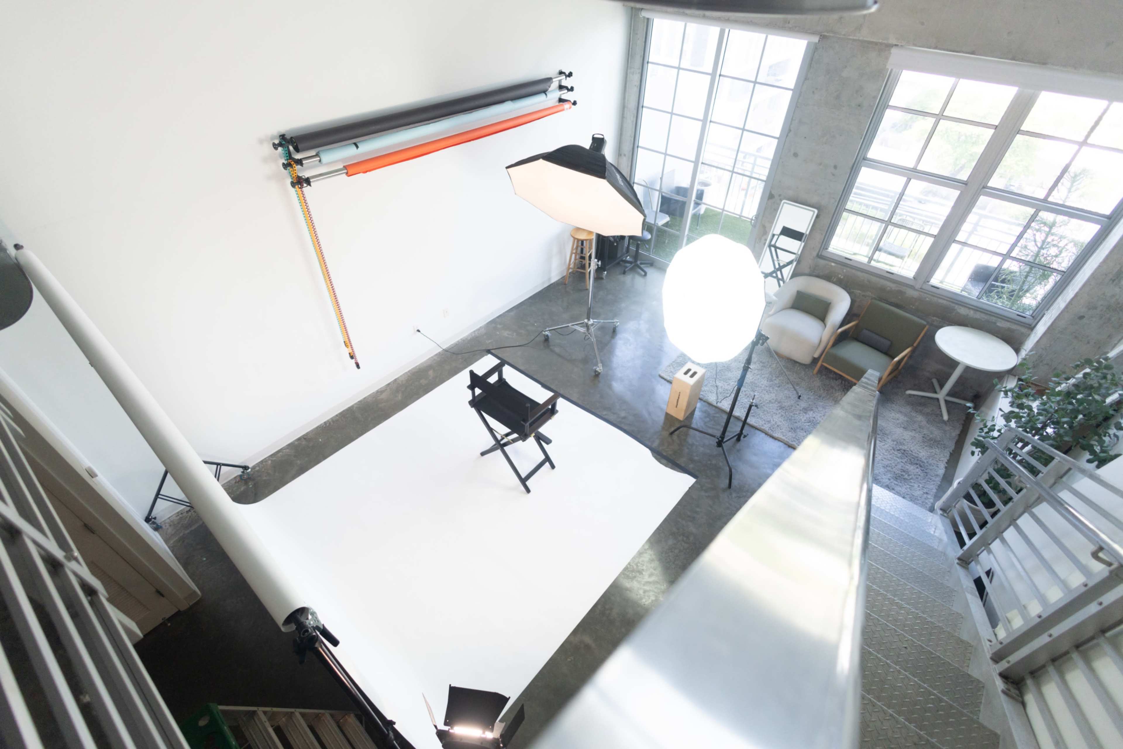 The image shows a well-lit photography studio with a white backdrop, equipment including lights and a director's chair, and a minimalistic setup featuring plants and furniture.