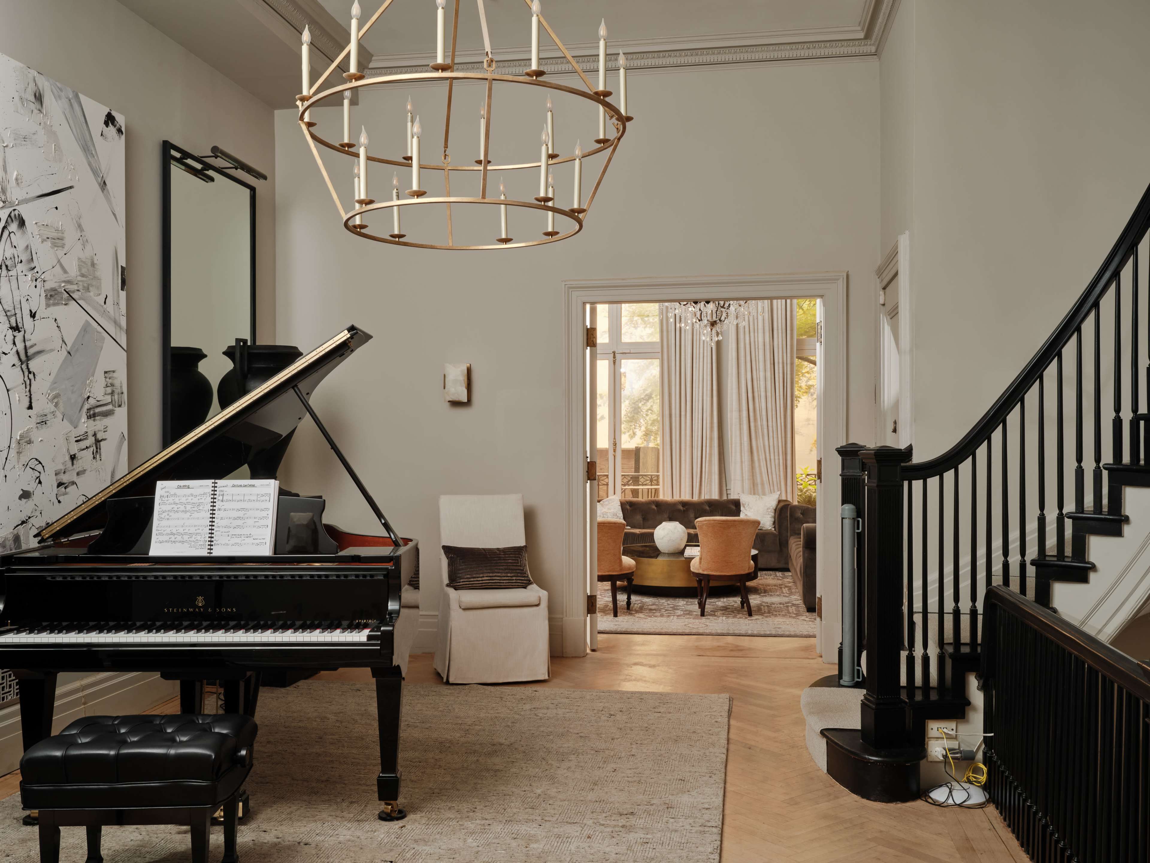 A grand piano with sheet music sits in a spacious entryway featuring a chandelier and a staircase, with an open living area visible through a doorway.