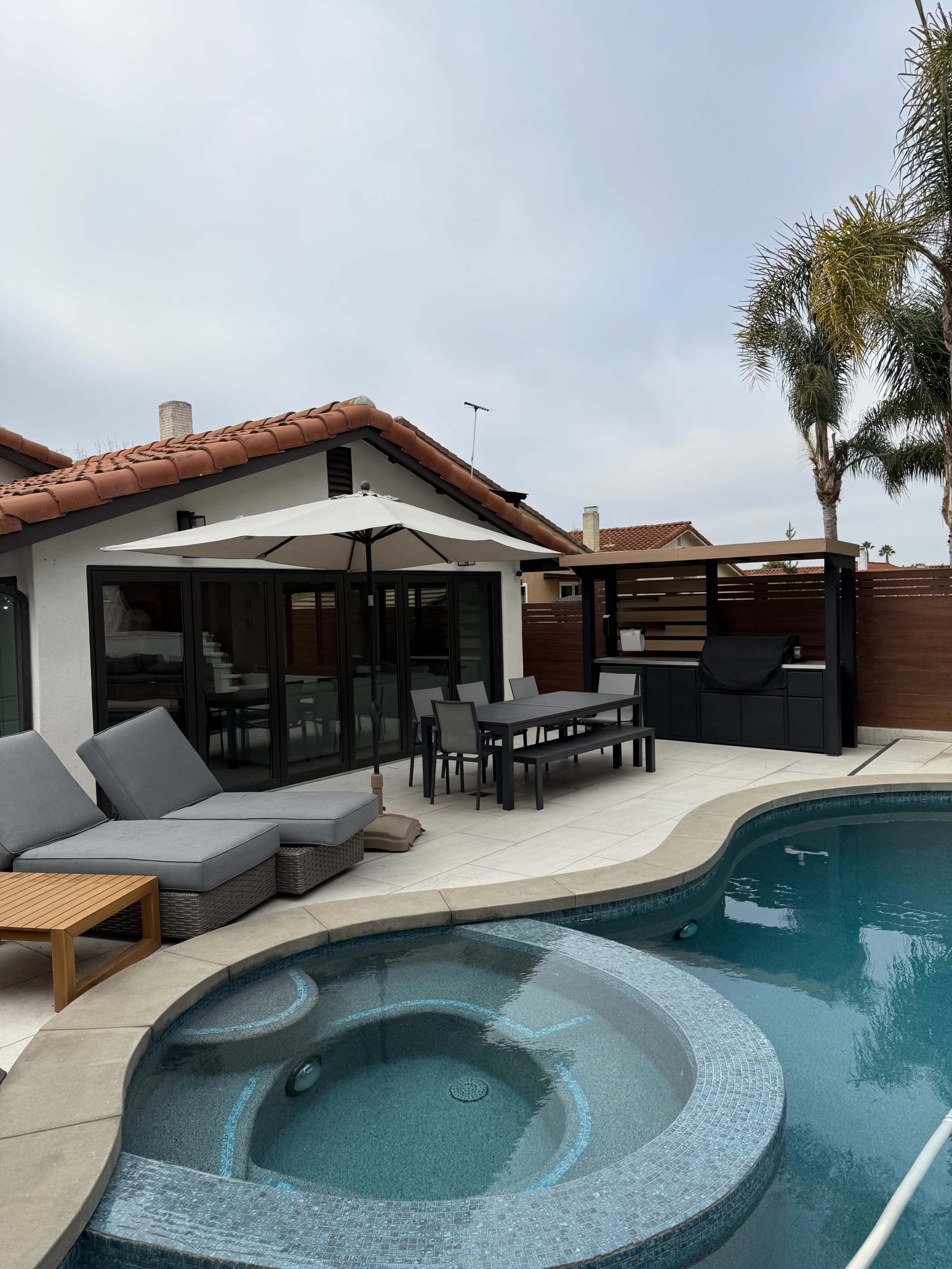 A backyard patio features a pool with a spa, lounge chairs, a dining table, and a covered bar area.