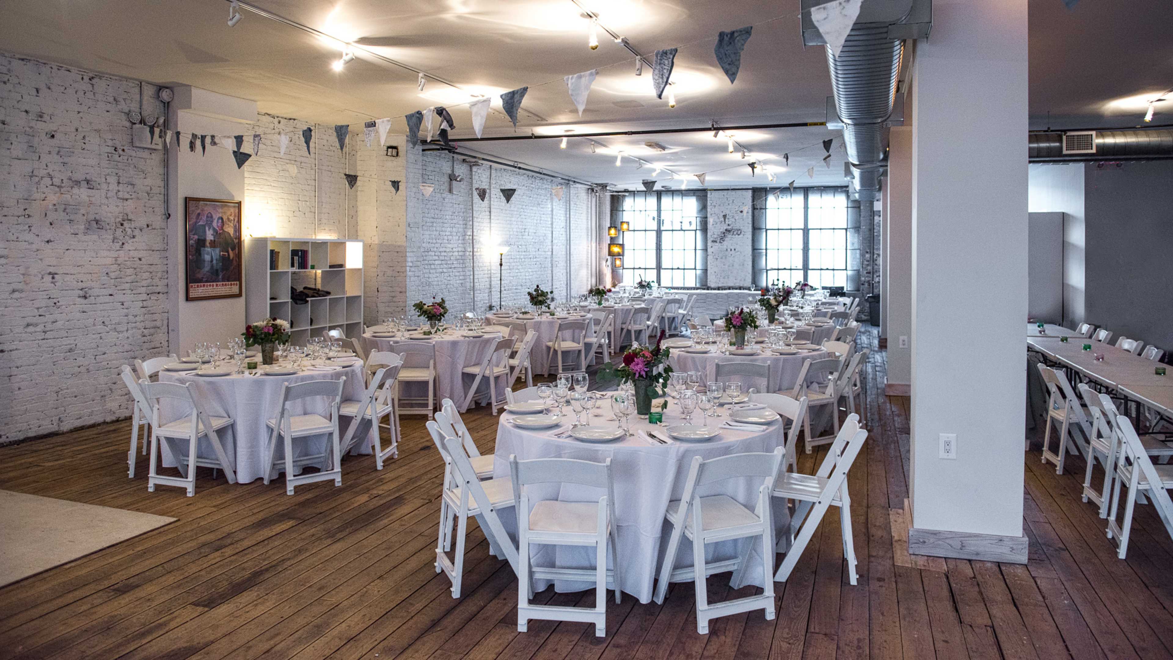 The image shows a spacious event room set for a formal gathering, featuring round tables covered with white tablecloths and decorated with floral centerpieces, along with bunting hanging from the ceiling.