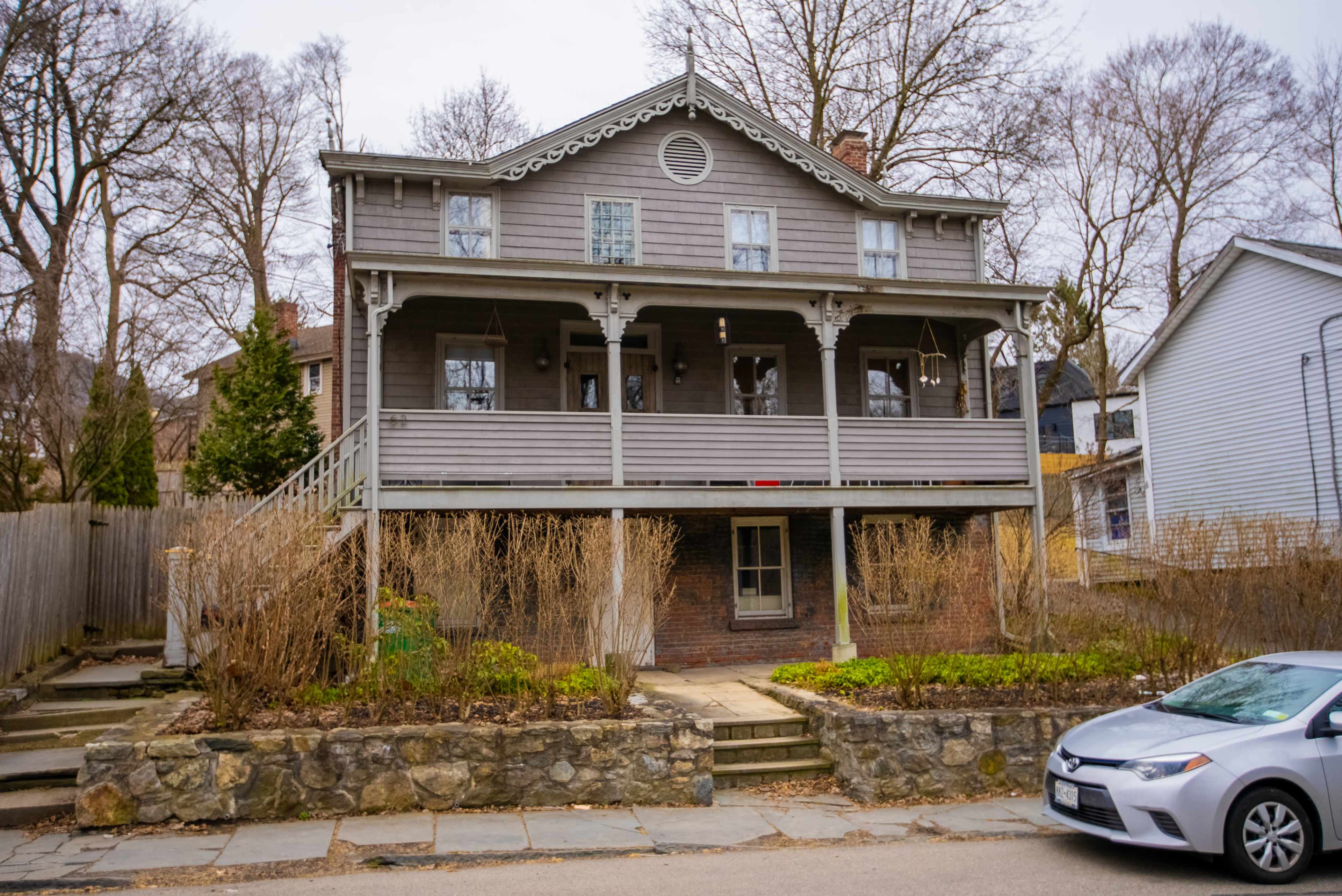 The image shows a two-story gray wooden house with a porch and stone pathway, surrounded by bare trees and adjacent to a parked gray car.
