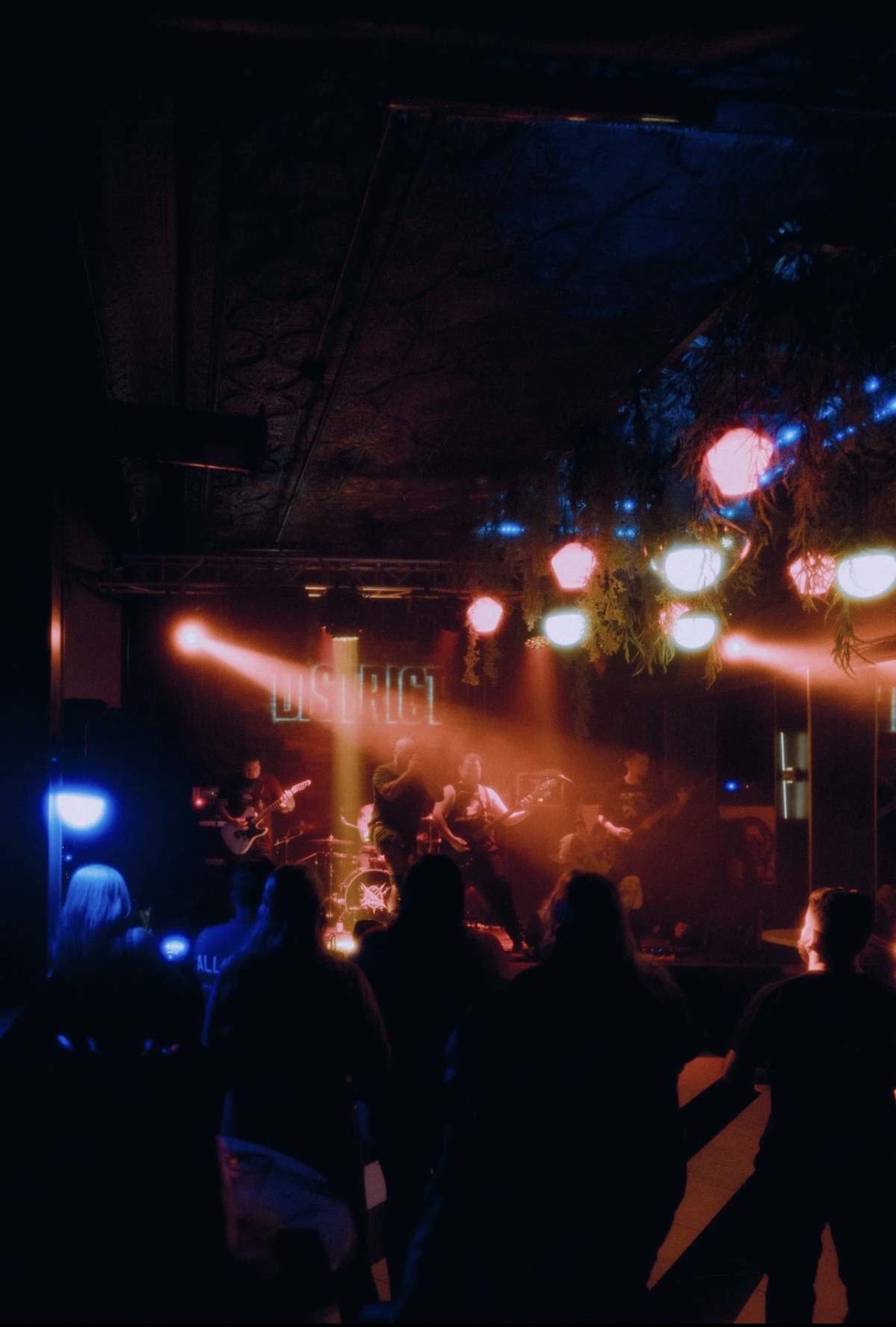 A live band performs on stage in a dimly lit venue while an audience watches from the foreground.