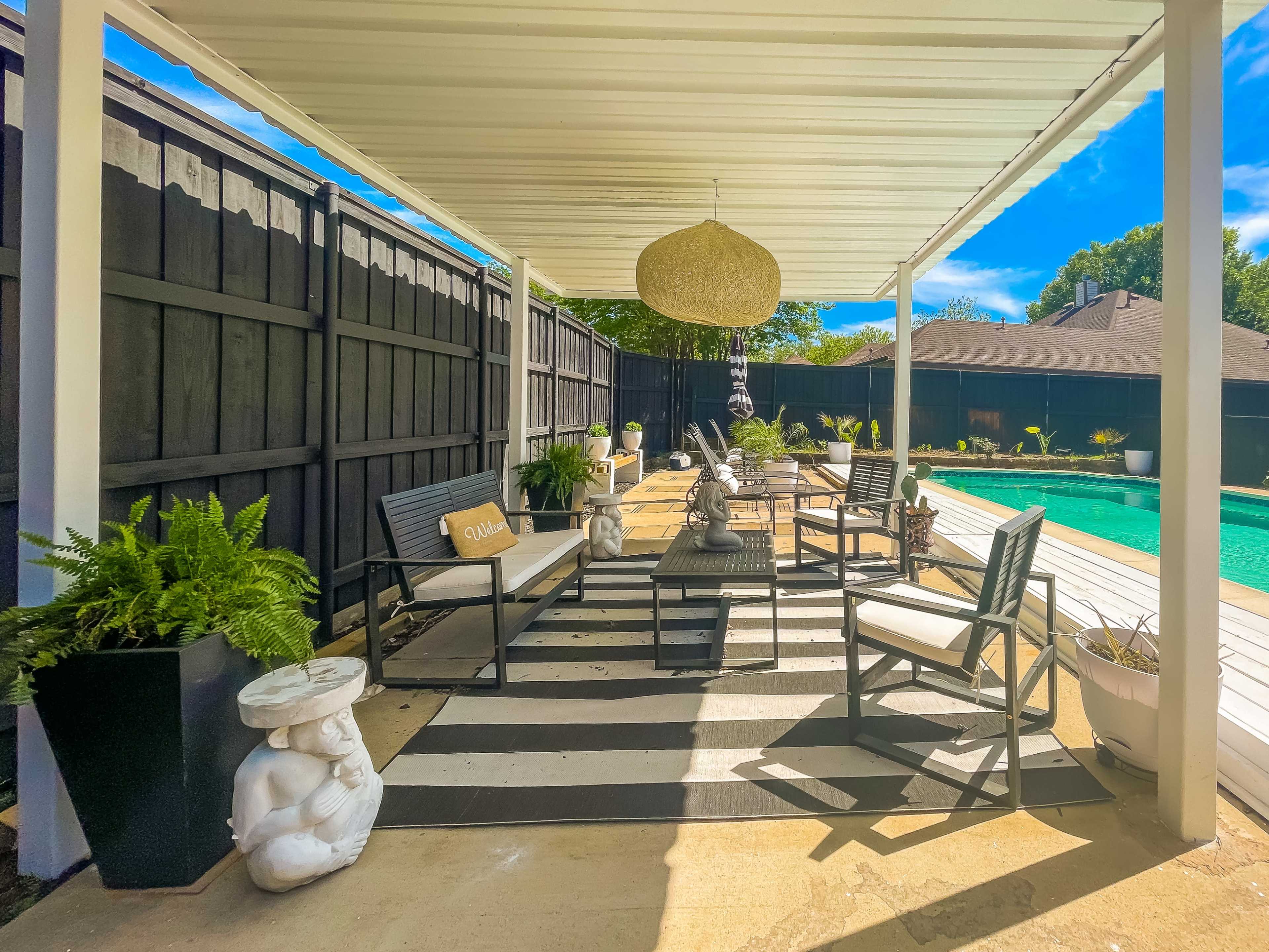 A covered outdoor seating area features black and white striped flooring, comfortable chairs, potted ferns, and decorative sculptures beside a pool.
