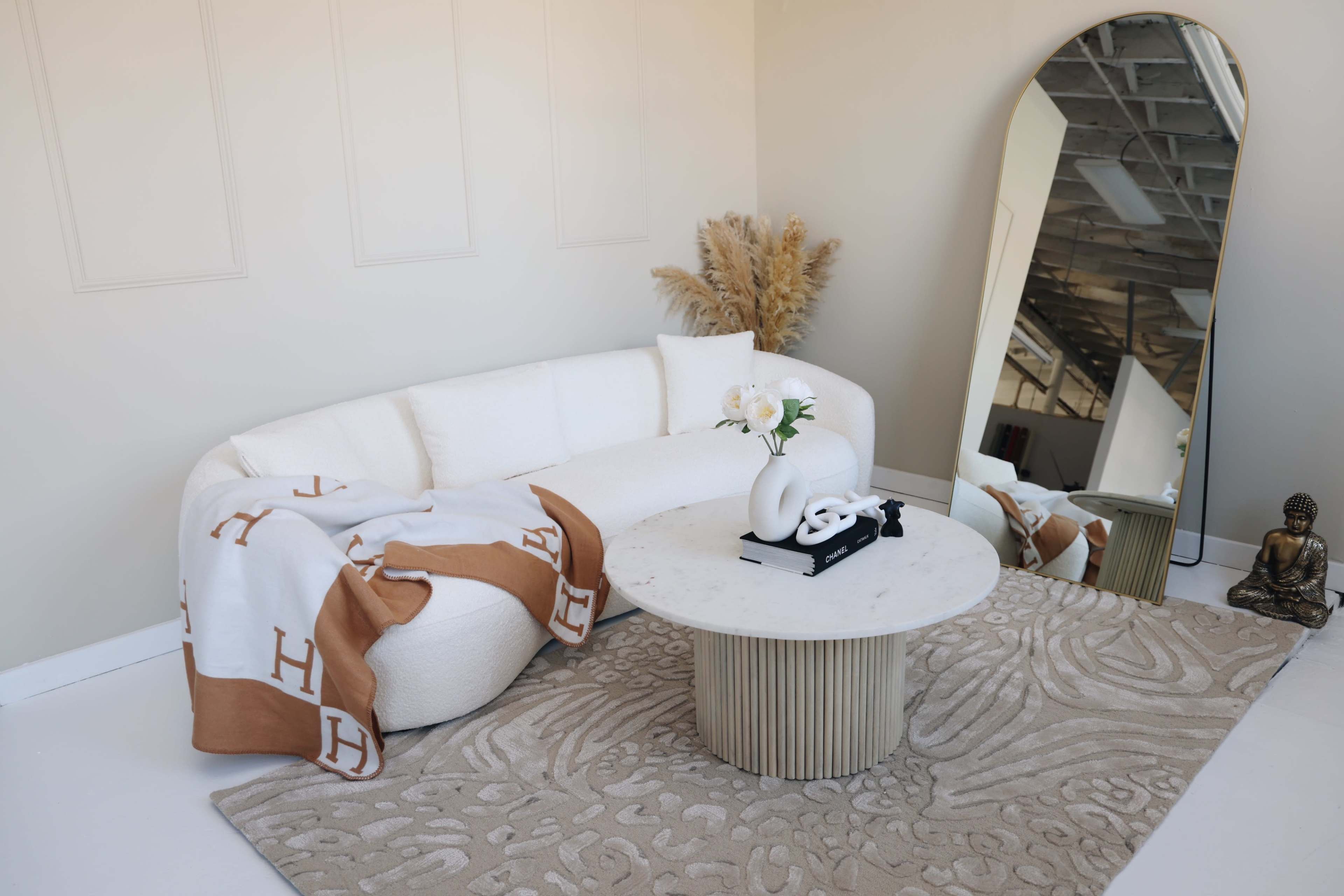 A modern, curved white sofa with a beige throw blanket sits near a large mirror and a round marble coffee table on a decorative rug in a minimalist room.