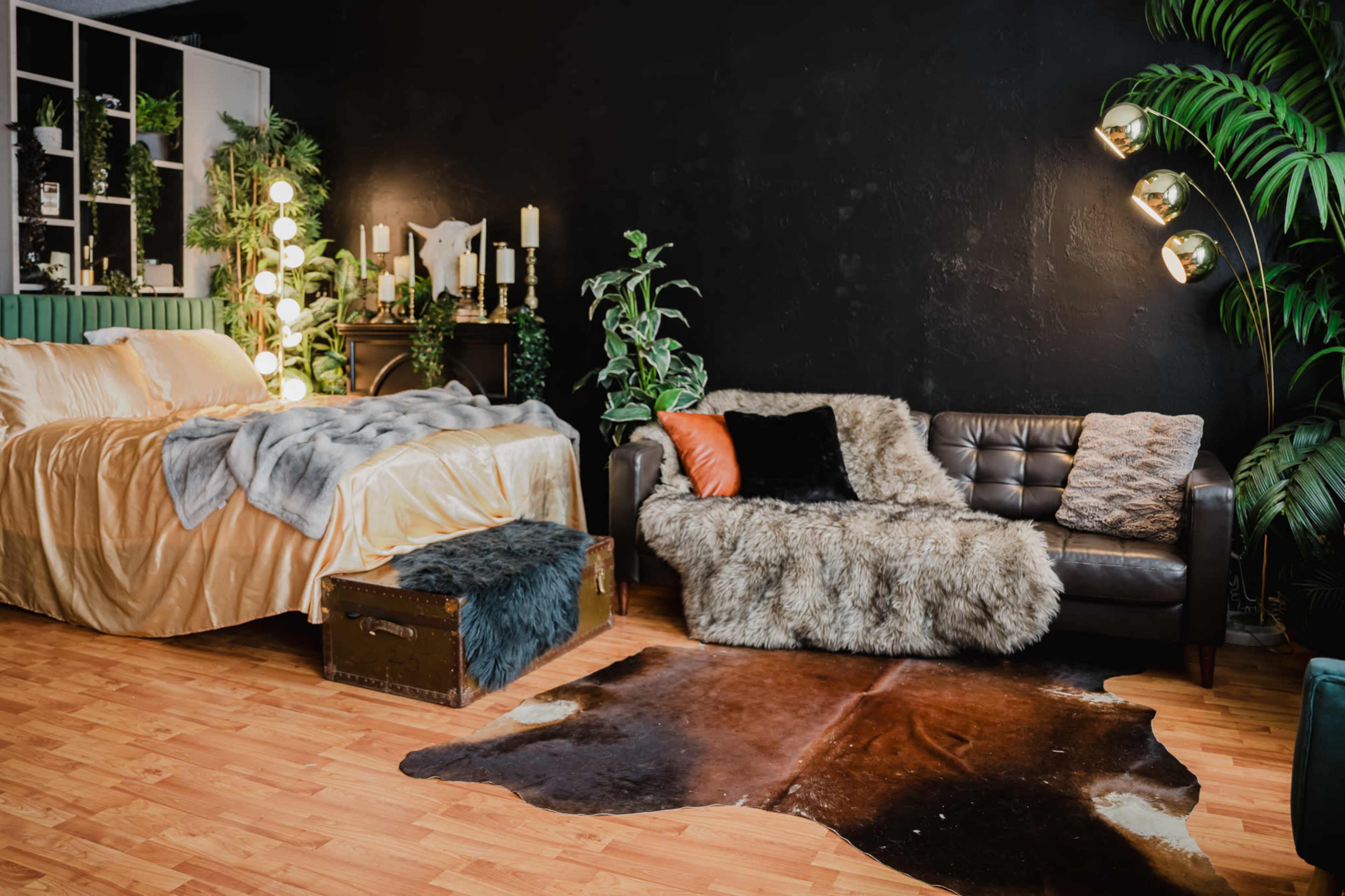 The scene features a modern bedroom with a large bed dressed in golden bedding, a cozy sofa draped with fur, and various plants accenting the dark walls.