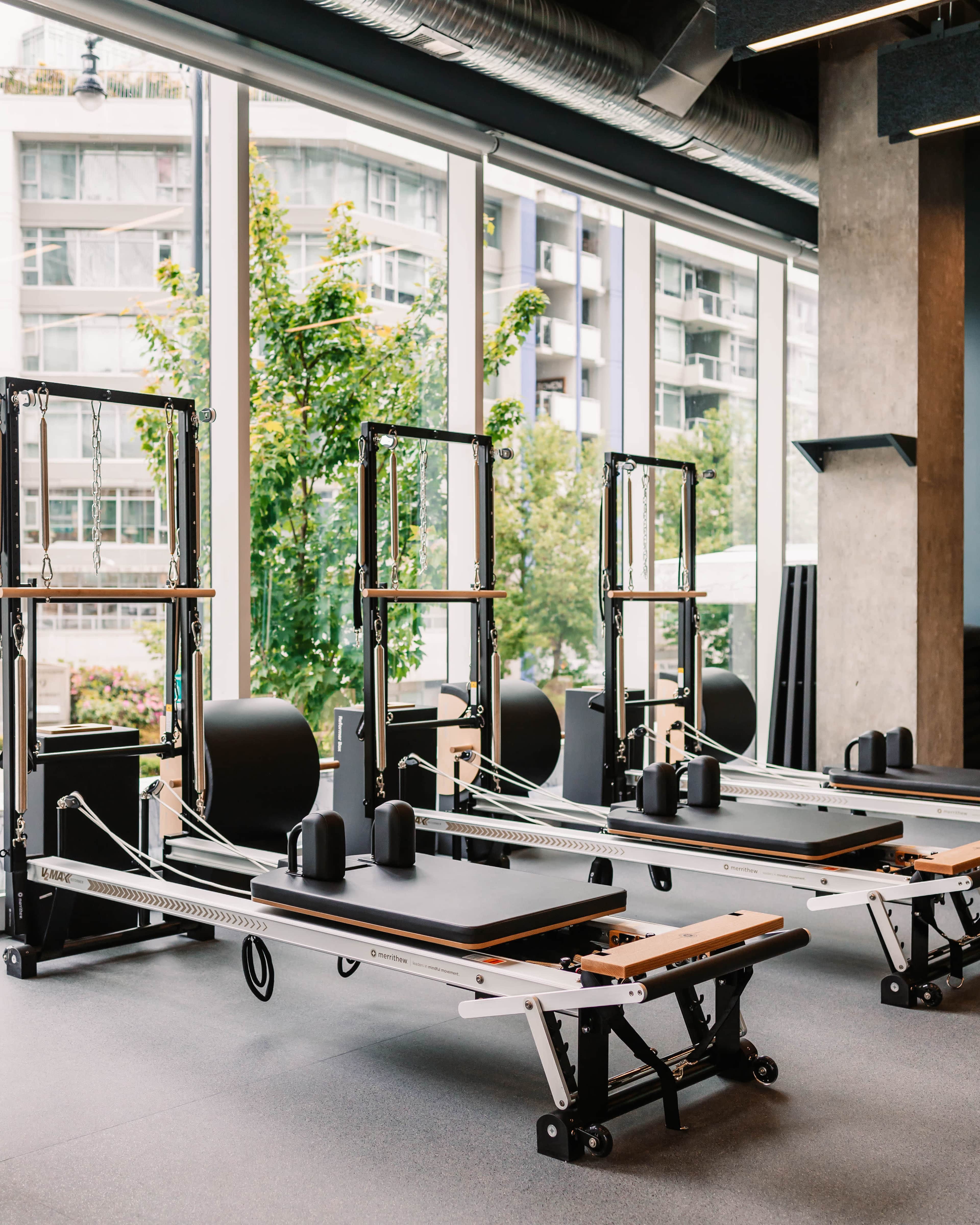 The image shows a fitness studio featuring multiple reformer Pilates machines arranged near large windows with views of greenery outside.